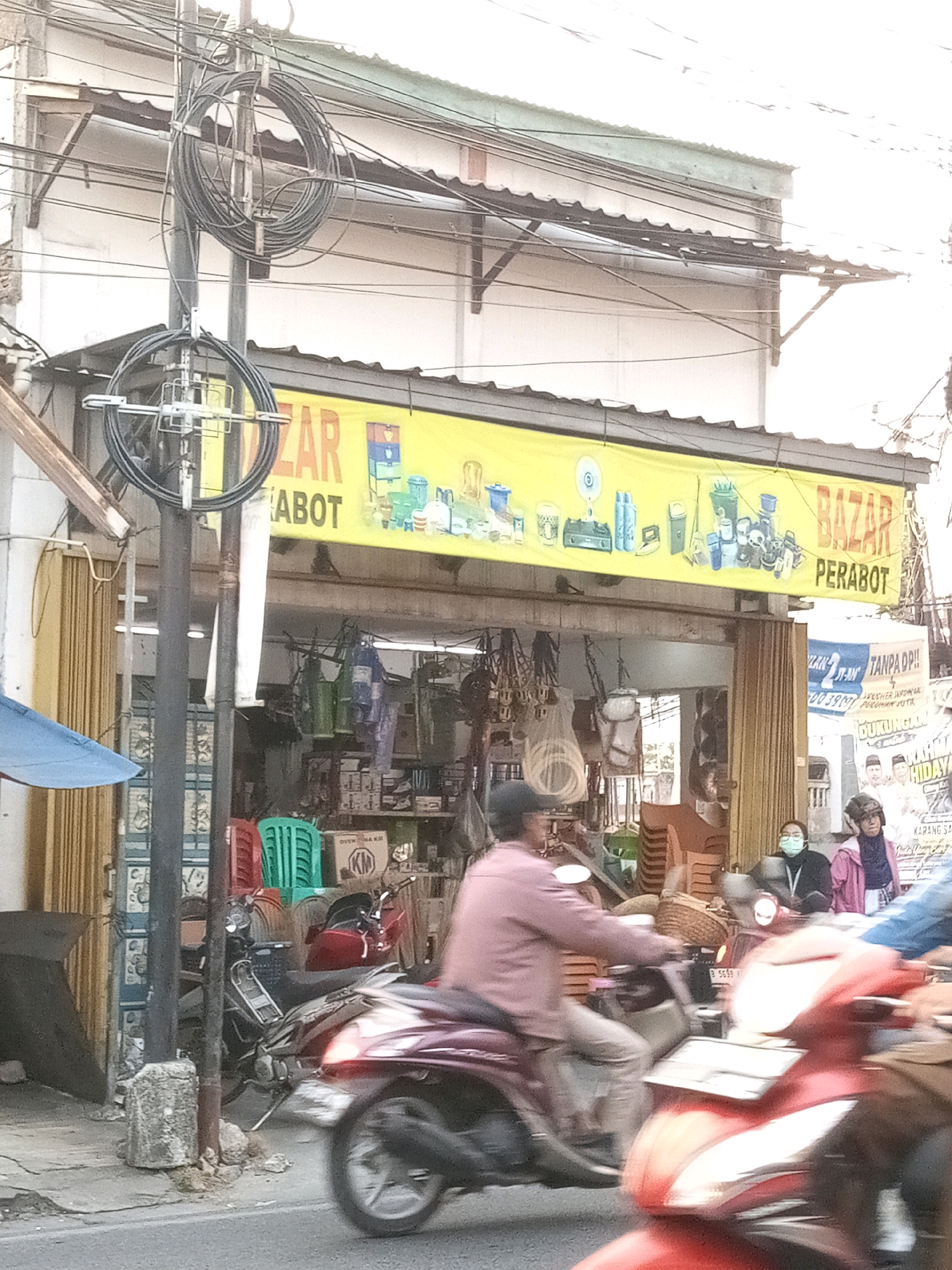 The image depicts a street scene in Karangsatria, Indonesia, featuring a "BAZAR PERABOT" (Furniture Bazaar) storefront. The storefront, characterized by a yellow banner, displays various household items. In the foreground, several motorcycles are visible, with one in motion, indicating traffic on the street. People are seen on and around the motorcycles. The environment includes a street, sidewalk, and the facade of the bazaar. Electrical wires are prominently strung across the top of the image, suggesting an urban setting. The overall impression is one of a bustling local market.