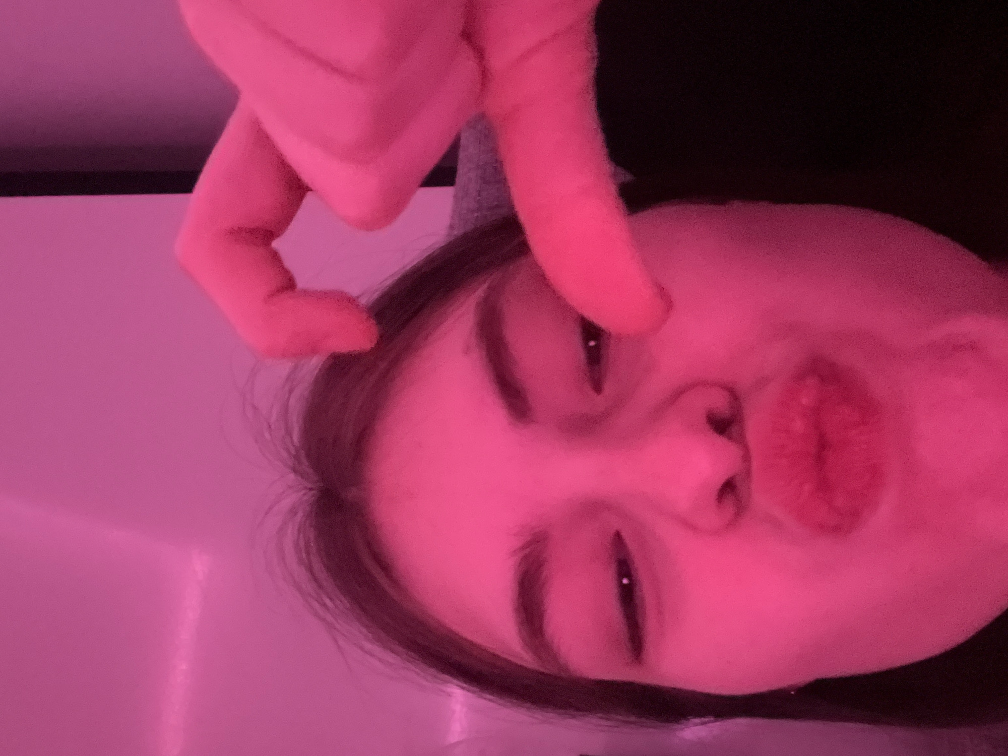 A close-up, tilted portrait shows a person with fair skin and dark hair, illuminated by a pink light source. The person's face is angled, with a slightly pursed expression. Above the face, a hand forms a shape, with fingers bent. The background is a mix of shadows and indistinct shapes, with a possible light source visible in the lower left corner. The scene is not recognizable as a location in Saint Petersburg, Russia.