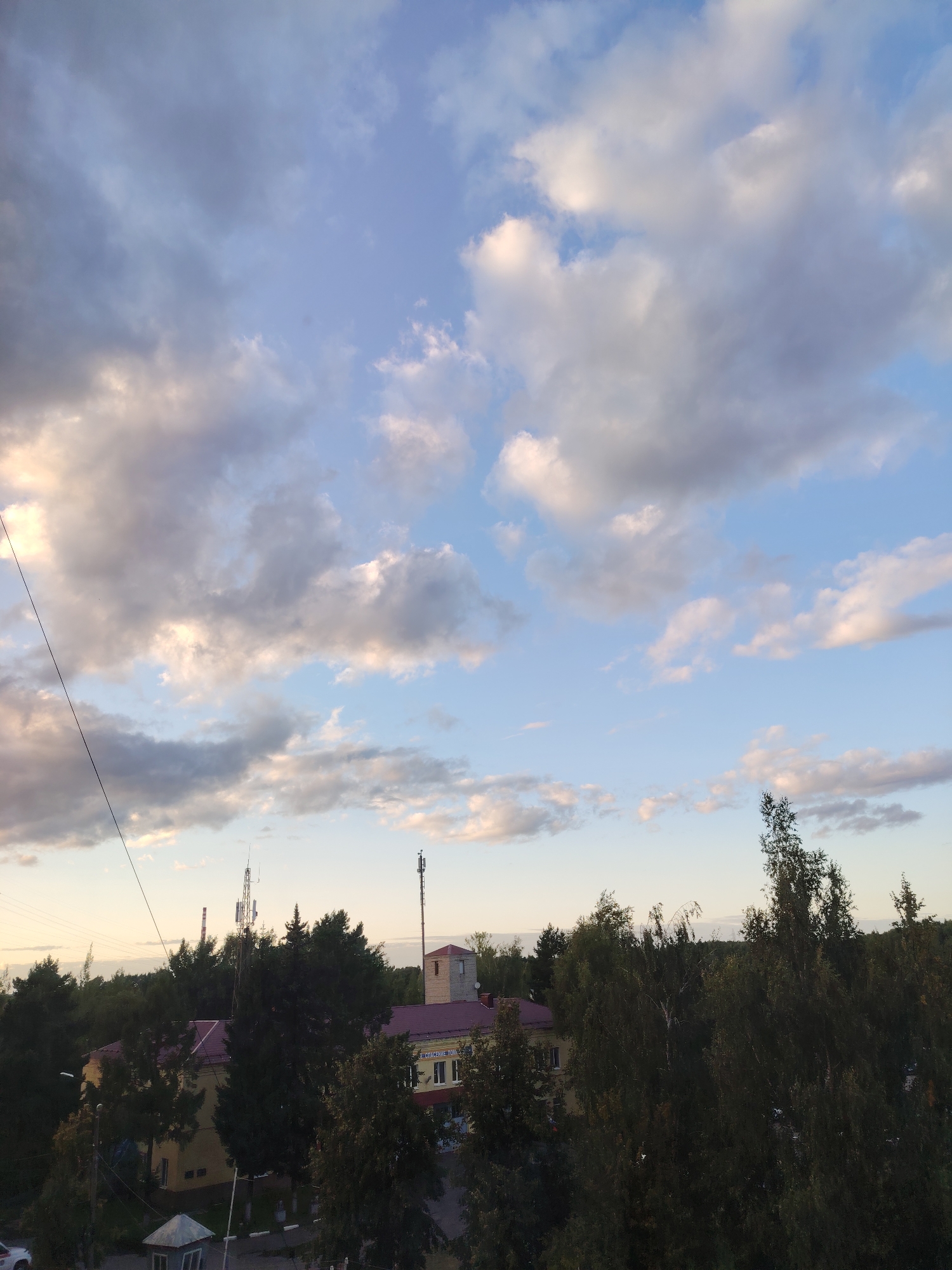 A high-angle, wide shot captures a view of the city of Konakovo, Russia, with a focus on the sky and a glimpse of buildings and trees below. The sky is filled with a mix of blue and white clouds, with some areas appearing darker and more dramatic, suggesting an approaching storm or the end of the day. In the middle ground, several trees form a dense line across the horizon, partially obscuring buildings and communication towers. One building, with a red roof, is clearly visible, and a sign that reads "СПАСЕНИЕ ПОЖАР" can be seen on its facade. The overall atmosphere is serene, with the natural beauty of the sky contrasting with the man-made structures.
