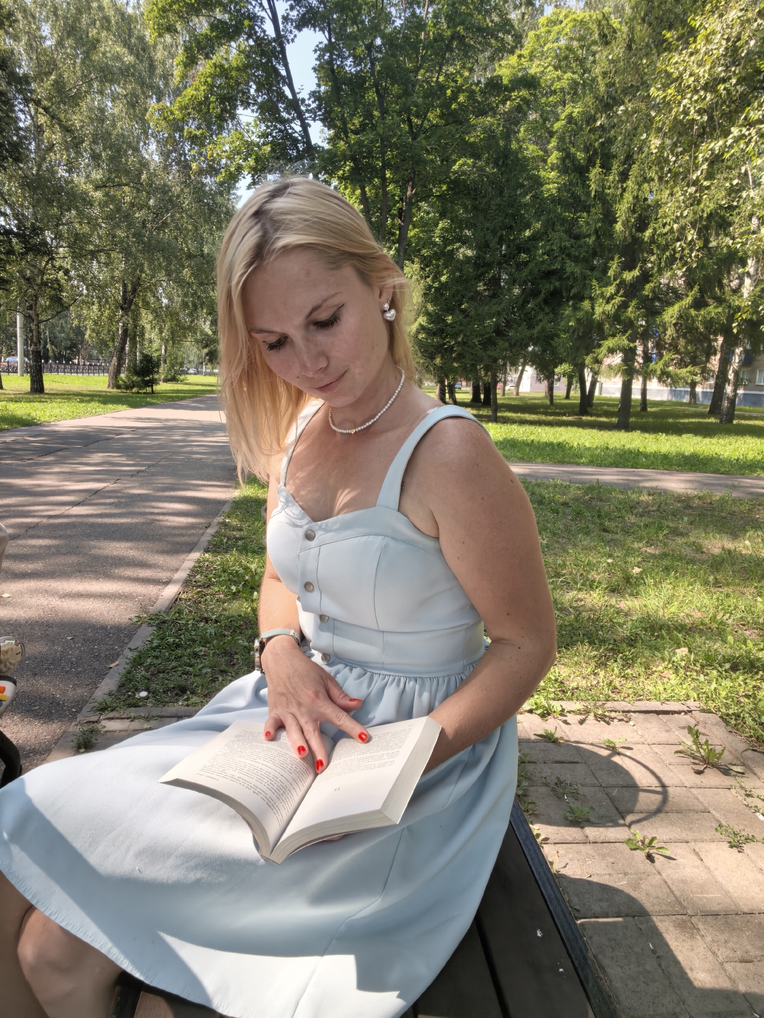 A woman with blonde hair is sitting on a park bench outdoors, reading a book. She is wearing a light blue sleeveless dress and a pearl necklace. Her fingernails are painted red. The park appears to be in a city, with trees and a paved path visible in the background. The lighting suggests it is daytime, possibly late morning or early afternoon. There are buildings visible in the distance through the trees.