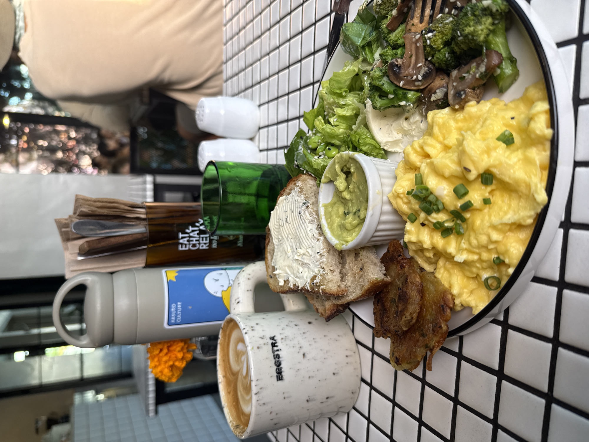 The image shows a meal on a table with a black and white tiled surface. A person is partially visible in the background, wearing a beige top. On the table, there is a plate of food with scrambled eggs topped with green onions, salad, sauteed mushrooms and broccoli, bread with cream cheese and guacamole, and fried fritters. Also on the table are a mug of latte with the word "EGGSTRA" written on it, a water bottle, cutlery, a small orange flower, and salt and pepper shakers. The writing "EAT CHAT RELAX" is visible on a glass near the cutlery. The scene appears to be at a cafe or restaurant, possibly in North Kuta, Indonesia.