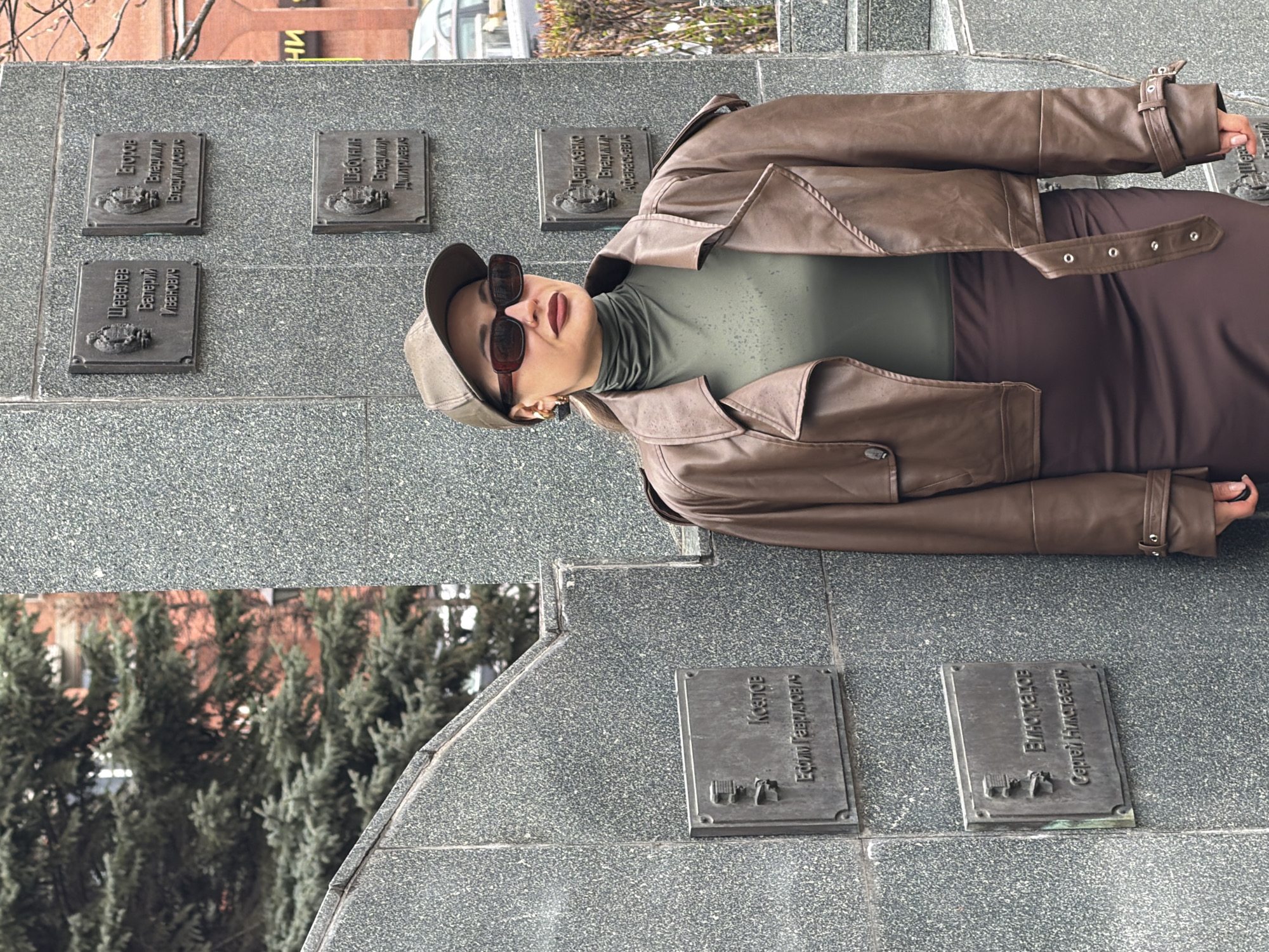 Here is a detailed description of the image:
**Scene:** The image appears to be taken outdoors, likely in the city of Krasnoyarsk, Russia, based on the Cyrillic inscriptions on the monument.
**Subjects:**
* **Person:** A woman is the main subject. She is wearing a brown leather jacket, a brownish dress, and a green turtleneck shirt. She has a brown cap on her head and is wearing brown sunglasses.
**Objects:**
* **Monument:** The woman is standing next to a gray monument with several metal plaques mounted on it. The plaques have Cyrillic text and some emblems on them.
* **Building:** In the background, a tall building is partially visible.
* **Trees:** There are evergreen trees or shrubs visible in the background.
* **Sunglasses**
* **Hat**
* **Jacket**
**Actions/Interactions:**
* The woman is posing for the photo, looking directly at the camera.
* The monument appears to be a memorial to individuals.
**Environment:**
* The weather seems cool, as the woman is dressed warmly.
* The location is urban, evidenced by the buildings.
* The environment is a public space with the monument and trees in view.