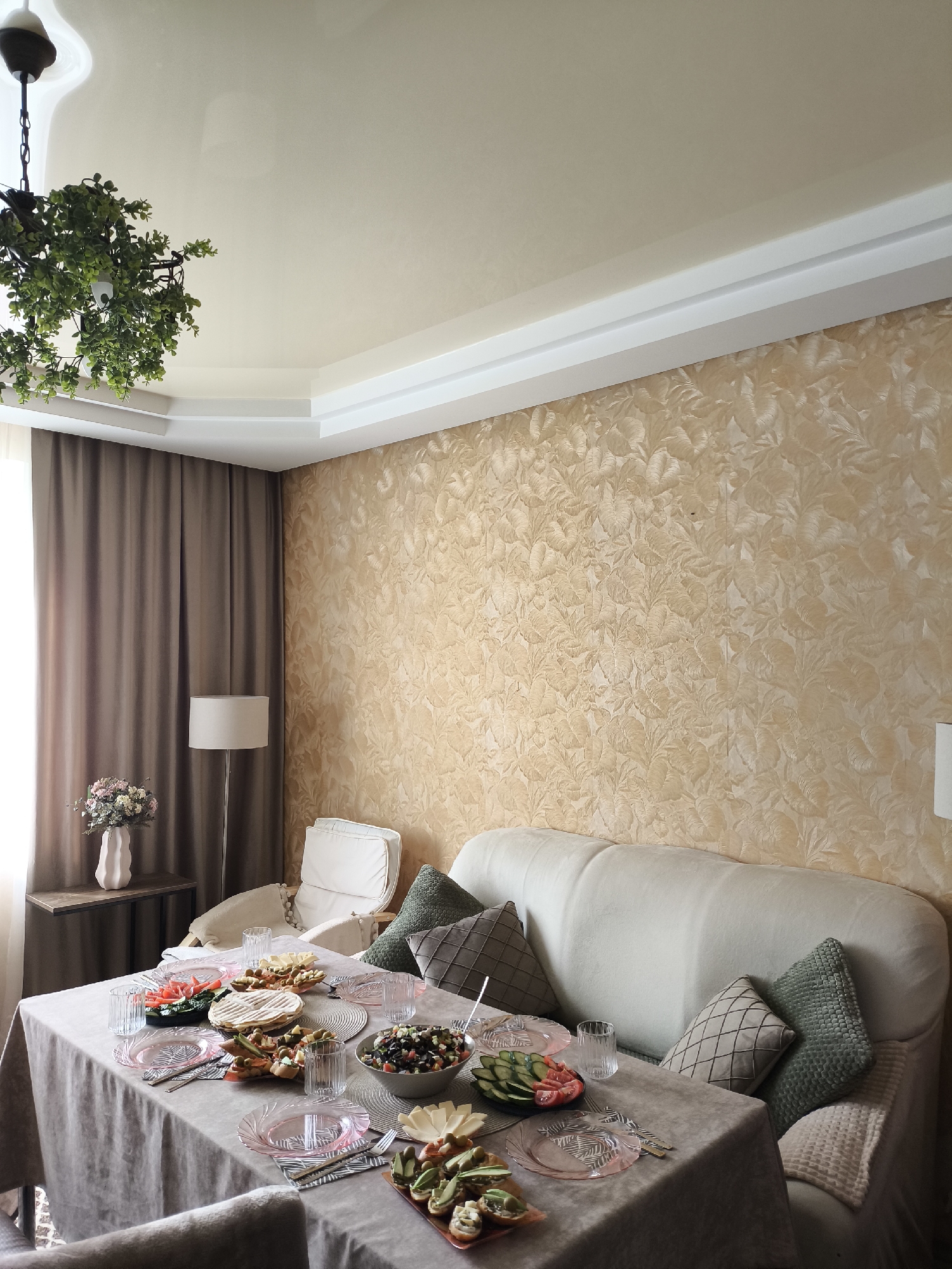 A medium shot captures a dining area in a home. A table covered with a grey tablecloth is laden with food and place settings, including pink plates, glasses, and cutlery. Dishes of salad, appetizers, and what appears to be flatbread are visible. Behind the table, a light-colored couch is adorned with decorative pillows in green and grey. To the left of the couch, a floor lamp with a beige shade stands next to a tall, brown curtain. A small side table holds a vase of flowers. The wall behind the couch features textured wallpaper with a leaf pattern in a light brown hue. The ceiling is white and multi-leveled, with a chandelier featuring artificial greenery hanging from it. The room appears to be well-lit, with natural light coming from the left.