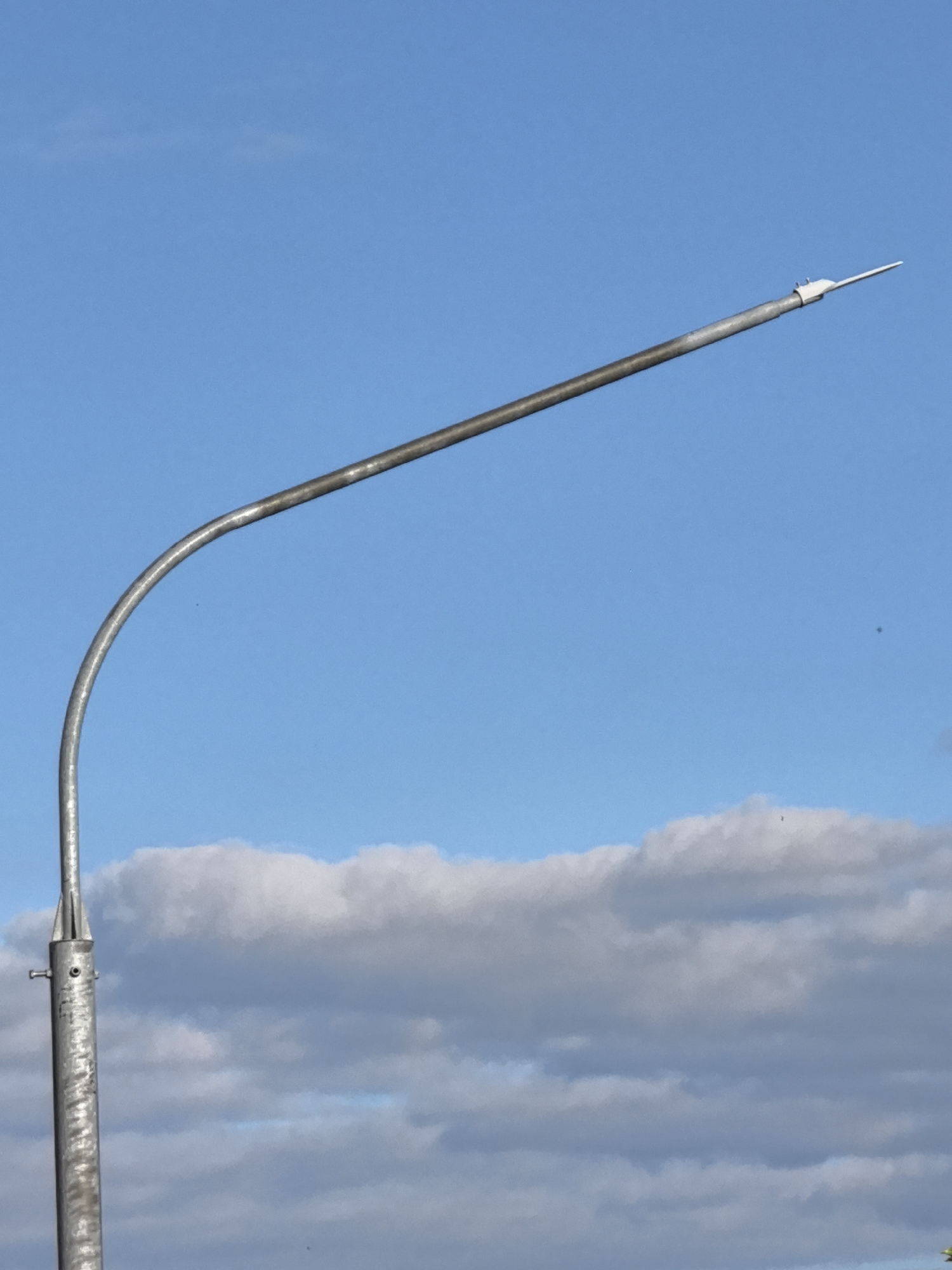 A tall, curved metal pole, likely a streetlamp, extends upwards against a bright blue sky. The pole is silver and appears to be made of galvanized steel, with visible texture and some imperfections. The curved section of the pole is a prominent feature, sweeping upwards and then straightening out to hold what appears to be a lightning rod at its tip. Below the curved section, the pole is mostly straight and vertical, with a cap-like fitting at its base.
In the lower portion of the image, fluffy white and gray clouds are scattered across the sky, suggesting a partly cloudy day. The clouds are illuminated, indicating it is daytime, likely with the sun overhead or slightly to the side. The overall scene is simple and uncluttered, focusing on the architectural element of the pole against the natural backdrop of the sky and clouds. There is no visible text in the image.