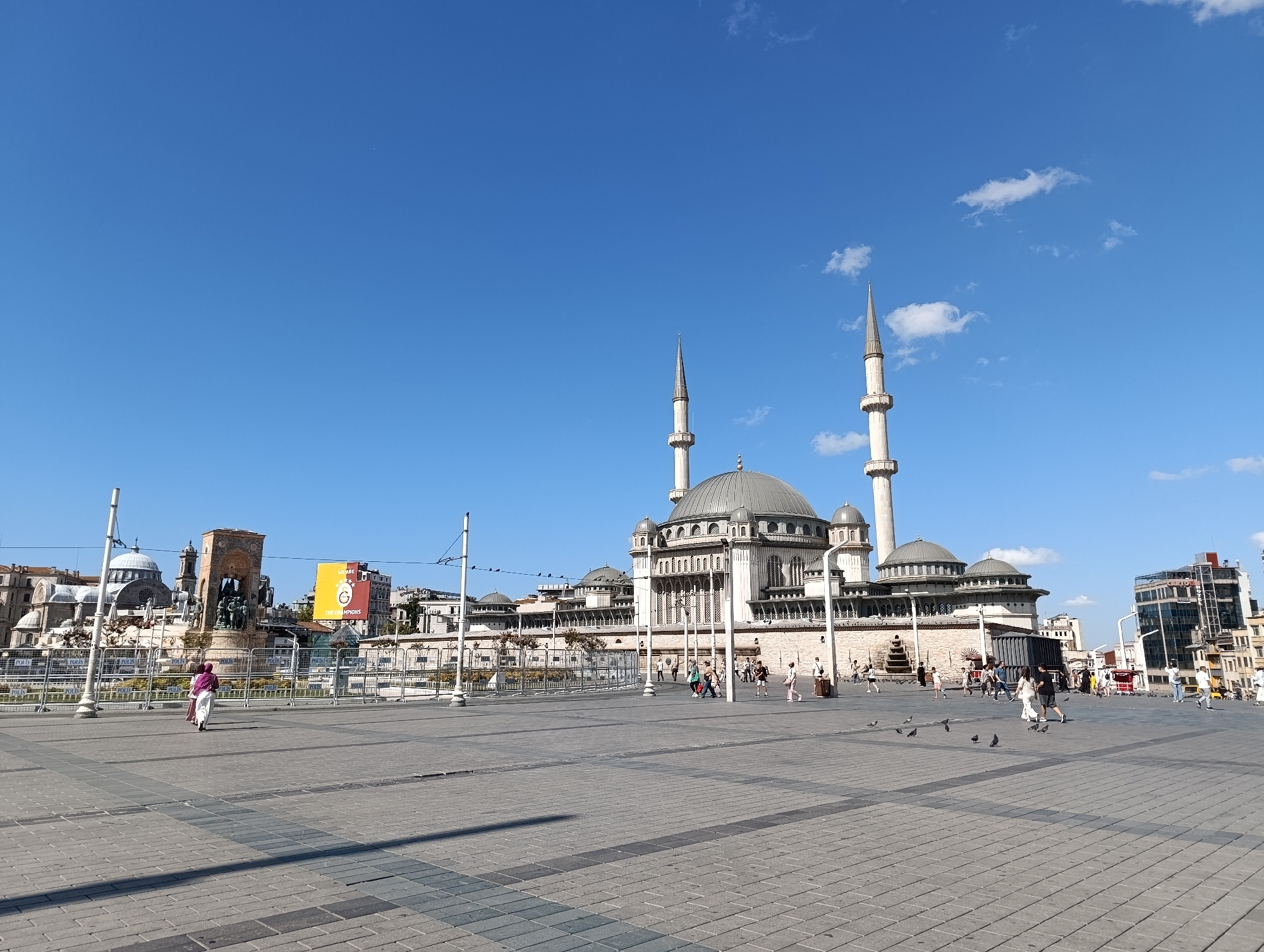 The image depicts a wide, paved plaza in front of the Kocatepe Mosque in the city of Beyoğlu, Turkey. The mosque is a large, imposing structure with a central dome, multiple smaller domes, and two tall minarets. The plaza is laid with patterned grey pavers and is largely empty except for a few scattered pigeons and a small group of people walking in the distance.
In the background, beyond the mosque, are several other buildings, including a modern glass-fronted structure. To the left of the mosque, a monument or statue is visible, surrounded by scaffolding and temporary fencing. A banner displaying a Galatasaray logo is hanging from one of the buildings. The sky is a clear, vibrant blue with a few wispy white clouds. The scene is illuminated by bright sunlight, casting distinct shadows on the plaza. A woman in a pink headscarf and white clothing walks away from the camera in the foreground.