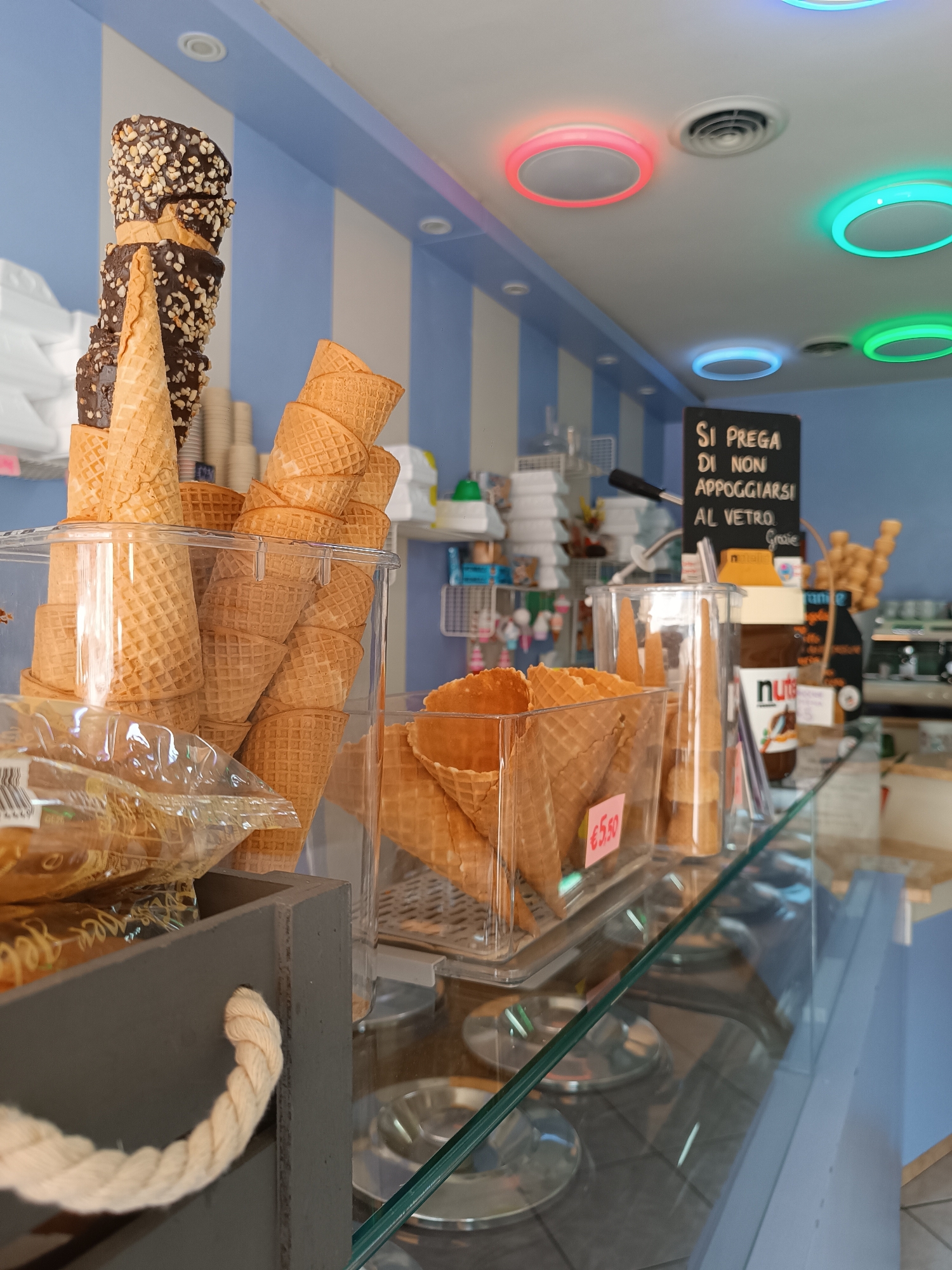 This image shows the interior of an ice cream shop in Casalmaggiore, Italy. The foreground features several clear plastic containers filled with waffle cones. To the left, a dark gray wooden crate with a thick rope handle is visible, containing bags of what appear to be waffle cone accessories. Behind the cones, a shelf holds various items, including a jar of Nutella and a coffee machine in the background. The ceiling is decorated with colorful circular lights, and a black sign with Italian text, which reads "SI PREGA DI NON APPOGGIARSI AL VETRO. Grazie," meaning "PLEASE DO NOT LEAN ON THE GLASS. Thank you," is prominently displayed. The walls are painted with alternating vertical stripes of blue and white. The overall scene depicts a typical ice cream parlor setting, with products neatly displayed for customers. No people are visible in the image.