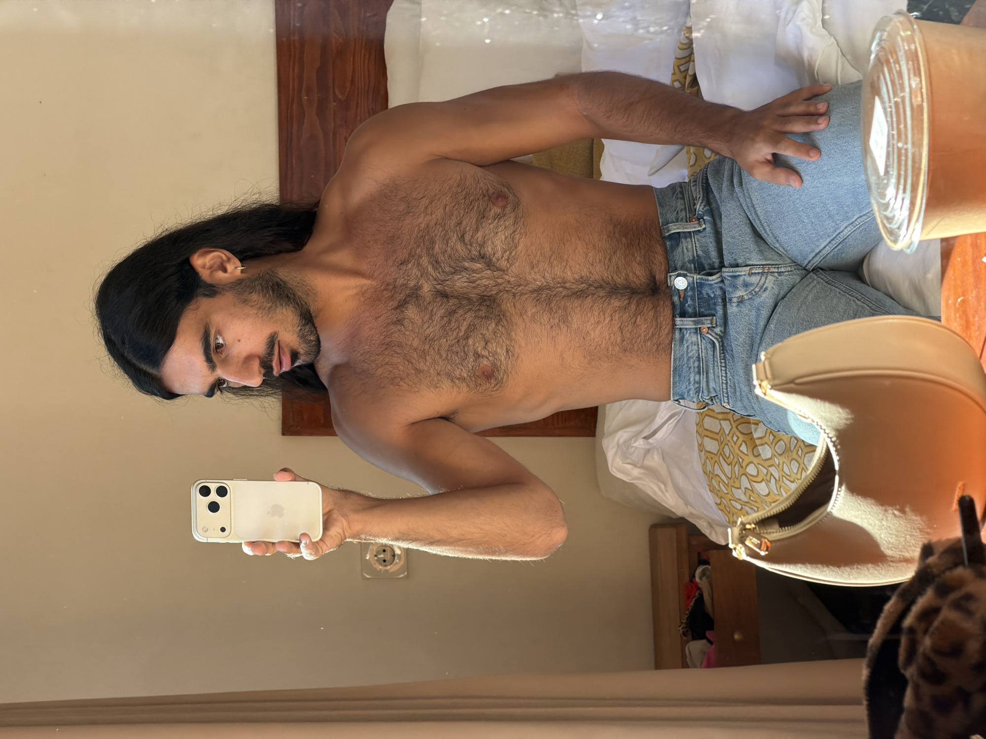 A shirtless person with long dark hair and a beard, wearing light blue jeans, takes a selfie in a mirror using a white iPhone. The subject is indoors, likely in a hotel room in North Kuta, Indonesia, given the decor and overall ambiance. Visible behind the subject are pillows, a wooden headboard, and a takeout container on the bed. A purse and stuffed animal are also reflected in the mirror.