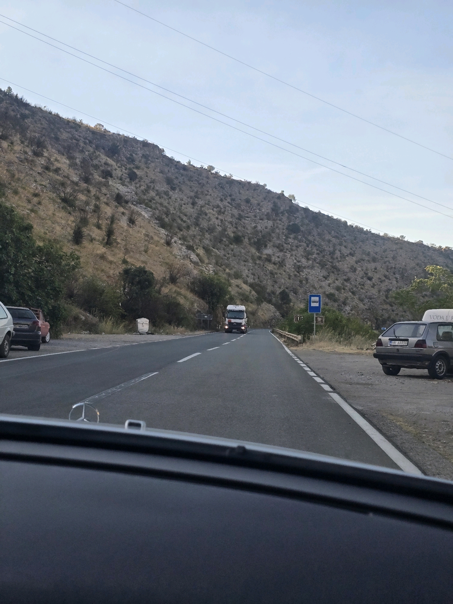 The image captures a roadside scene in Podgorica, Montenegro, characterized by a paved road stretching through a mountainous landscape under a clear sky. The road has distinct white lane markings. Several vehicles are present: a white truck is seen driving away from the viewer, and parked on the right side of the road is a silver hatchback. The terrain is rocky and covered with sparse vegetation. Overhead, power lines traverse the sky. A bus stop sign is visible on the right side of the road. The perspective suggests the photo was taken from inside another vehicle, looking forward.