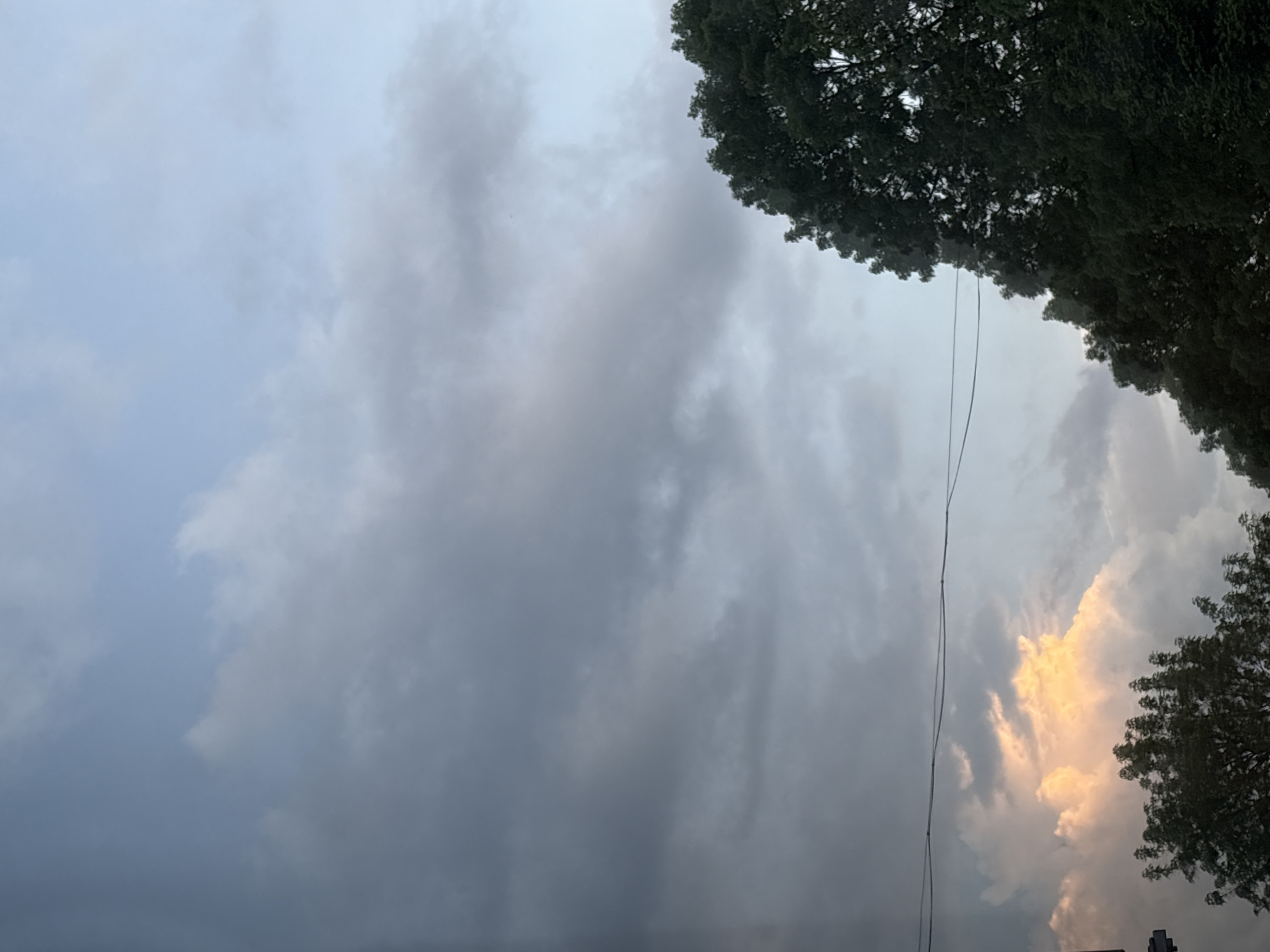 The image captures a sky view in Panagyurishte, Bulgaria. The scene is dominated by a sky filled with clouds, with a mix of dark, ominous gray clouds on the left and lighter, sunlit clouds on the right. The sun illuminates the clouds in the lower right. Some tree branches are visible at the top right and bottom right. Overhead power lines run down the center of the shot. The overall impression is of a cloudy day in the city.