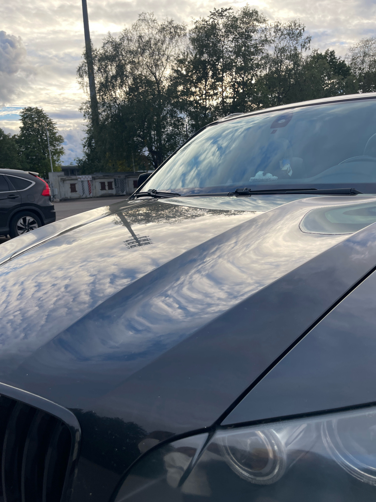 The image shows a dark colored car, likely an SUV, parked outdoors. The car's hood is highly reflective and shows a distorted reflection of the sky, which is partly cloudy, and some trees. A tall pole or pylon is also visible in the reflection and in the background. In the background, another dark colored SUV is parked. There are some buildings in the background as well, with what appears to be a red and white striped pattern. The scene suggests a parking lot or an area near a road in an urban or suburban environment. The lighting indicates it is daytime, possibly late afternoon or early morning given the sky's appearance and the angle of the light. No people are visible in the image. The location is in the city of Tallinn, Estonia.