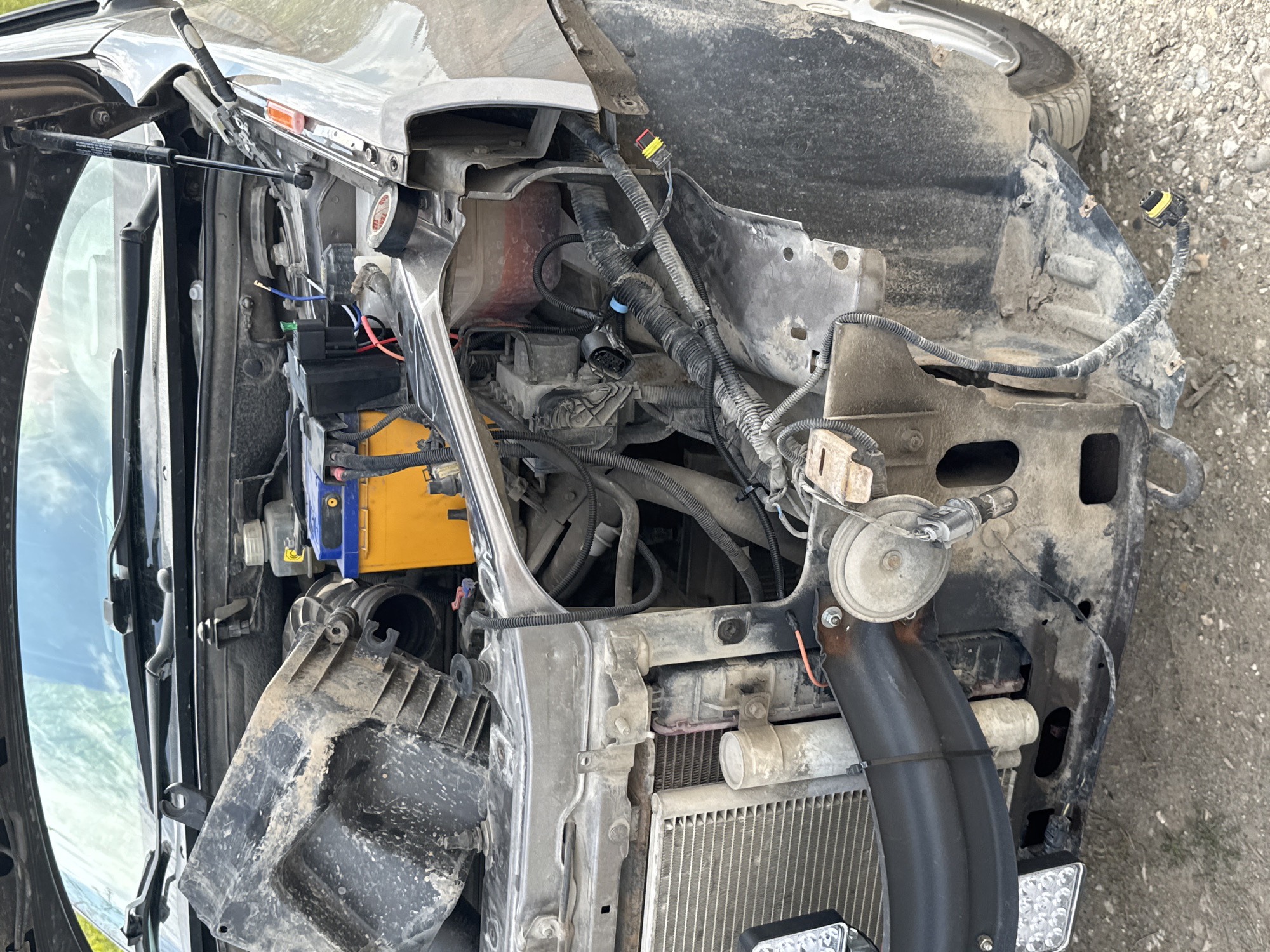 The image depicts the engine compartment of a damaged vehicle. The front end is heavily damaged, with crumpled metal and exposed wiring. A yellow and blue battery is visible on the left side. A radiator is located at the bottom. The vehicle is covered in dirt and debris. The location is unknown. There are no visible people.