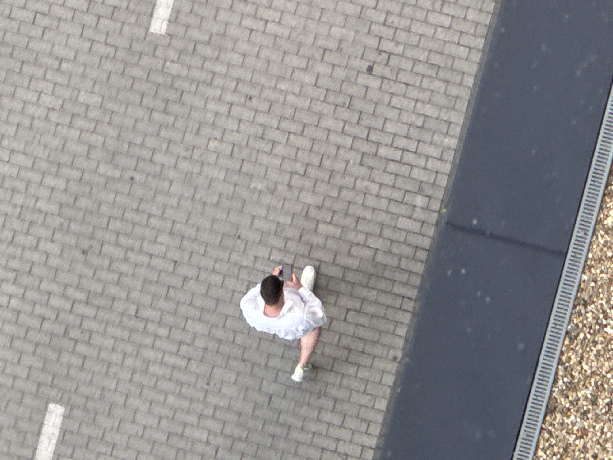 An overhead shot shows a man standing on a brick walkway, using a mobile phone. He is wearing a white shirt and white shoes. To his right, there is a dark-colored strip of pavement or road, followed by a drainage ditch and a gravel area. The scene is located in an urban setting. Based on context clues, it is potentially in Belgrade, Serbia.