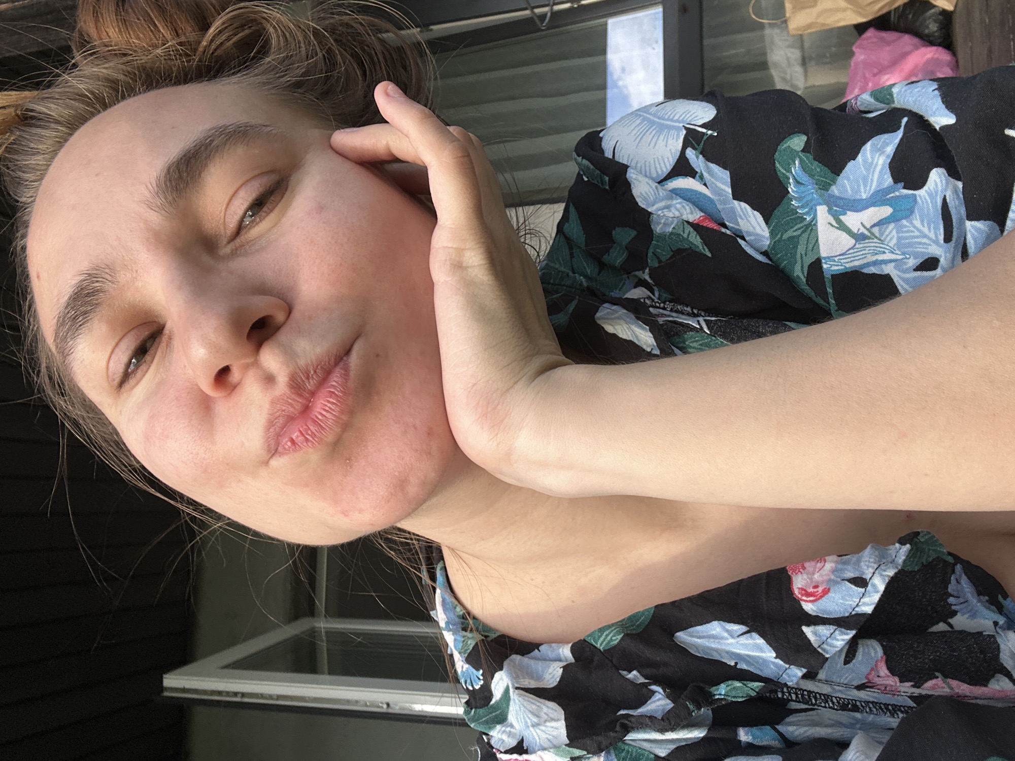 A fair-skinned person with brown hair pulled up in a bun is positioned at an angle, with their face tilted towards the camera. Their hand is placed on their cheek. They are wearing a black floral patterned shirt with shades of blue, green, and white. A window is visible in the background. There is no indication of the city of Kharnavy, Russia, in the image.