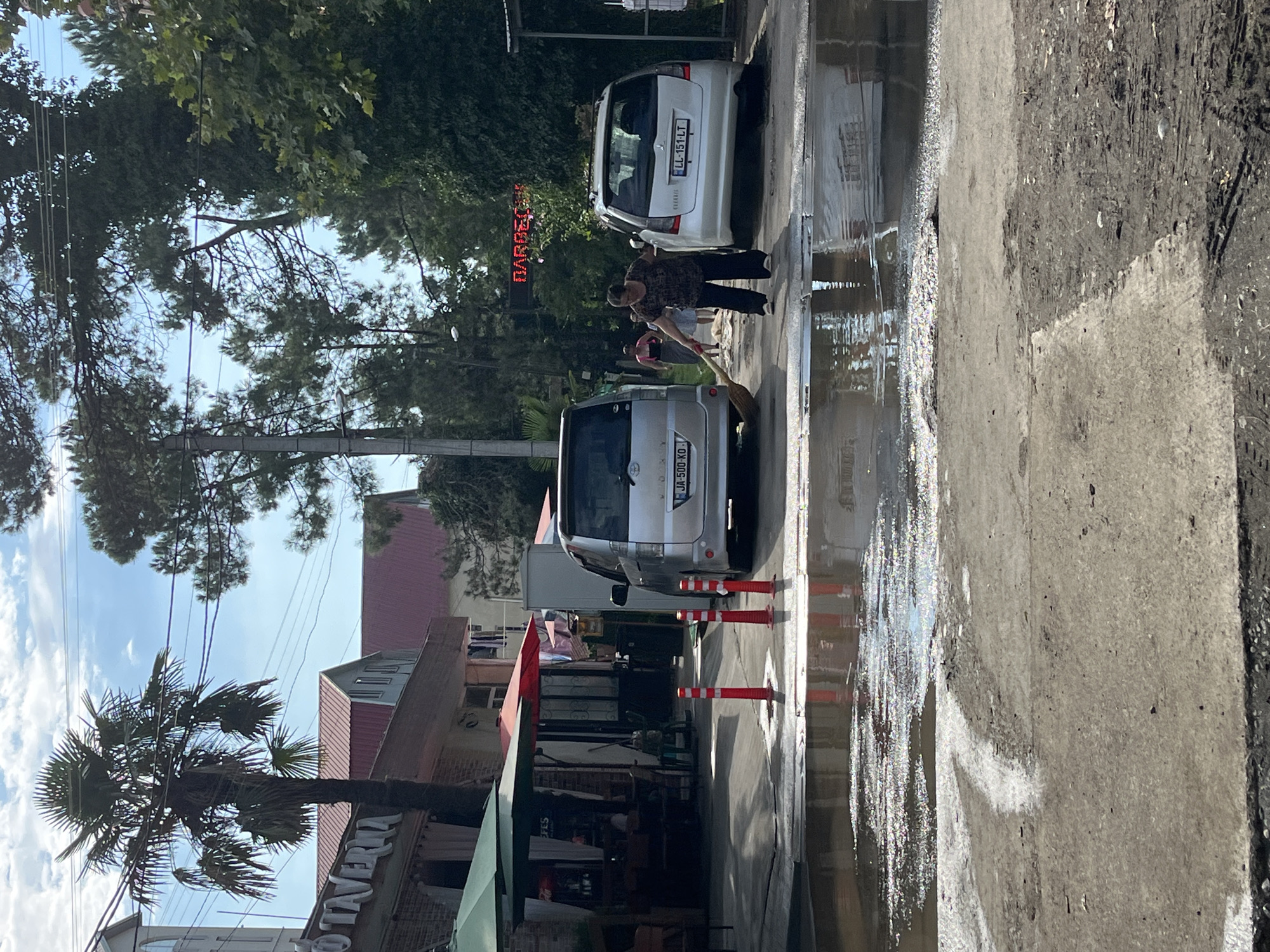 In Ureki, Georgia, a woman sweeps water from a flooded asphalt area in front of a building with a red-tiled roof and a sign reading "TAVERNA." Two silver minivans are parked nearby, one on the left with license plate "JA 500 KO" and another on the right with license plate "LL 151 LT." Several red traffic cones are placed in the flooded area. In the background, a large palm tree stands tall against a bright blue sky with scattered clouds. Trees and electrical wires are visible. A red LED sign displaying the word "BARBECUE" is also present in the background.