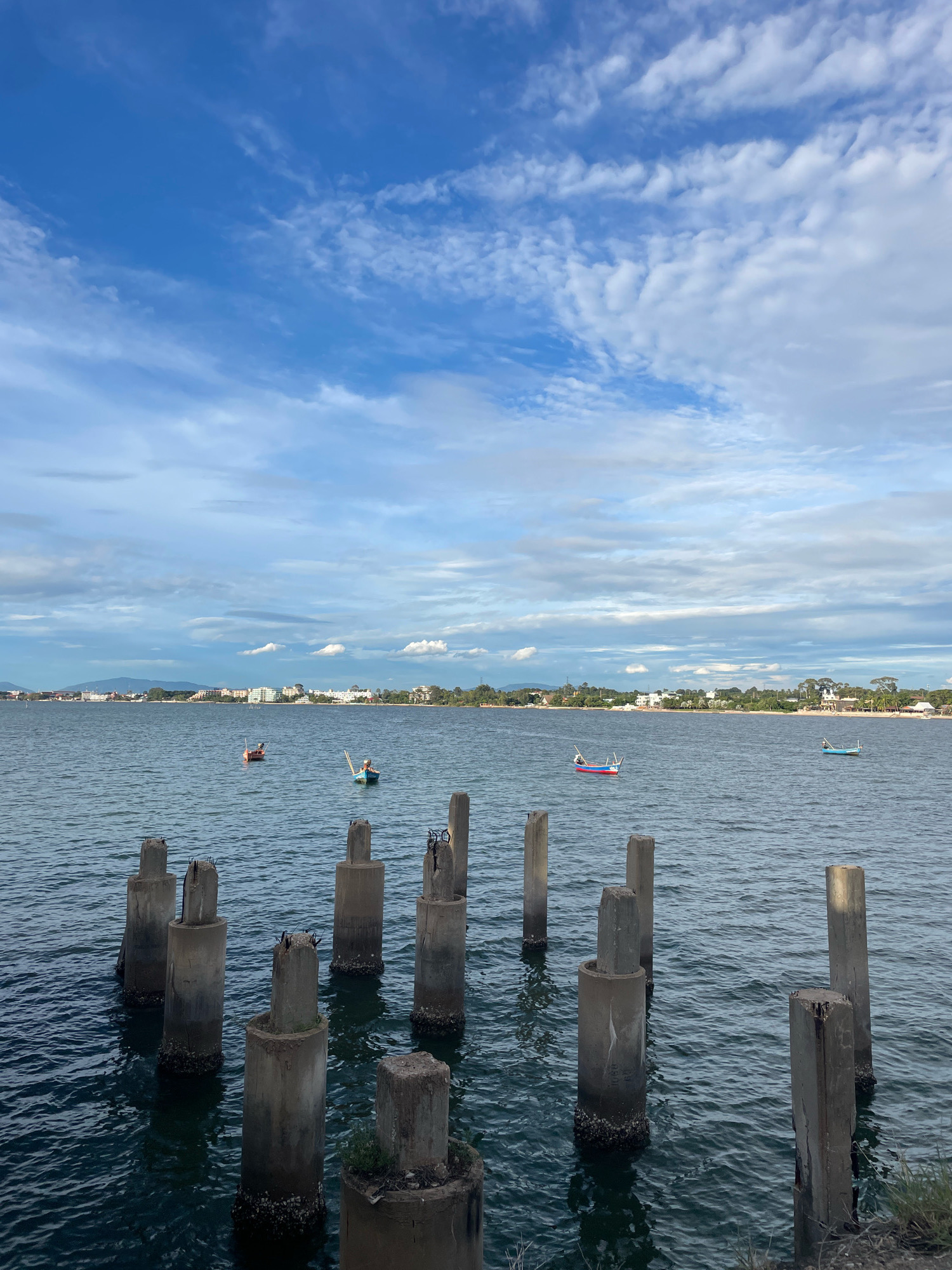 A photograph taken from the shore of the sea in Pattaya City, Thailand, shows a tranquil scene with a line of concrete pillars rising from the water in the foreground. Beyond the pillars, the calm sea stretches towards a distant coastline dotted with buildings and lush greenery. Several small fishing boats are visible in the water, some with occupants. The sky above is a clear blue, adorned with scattered white clouds, creating a serene atmosphere. The lighting suggests it is either early morning or late afternoon.