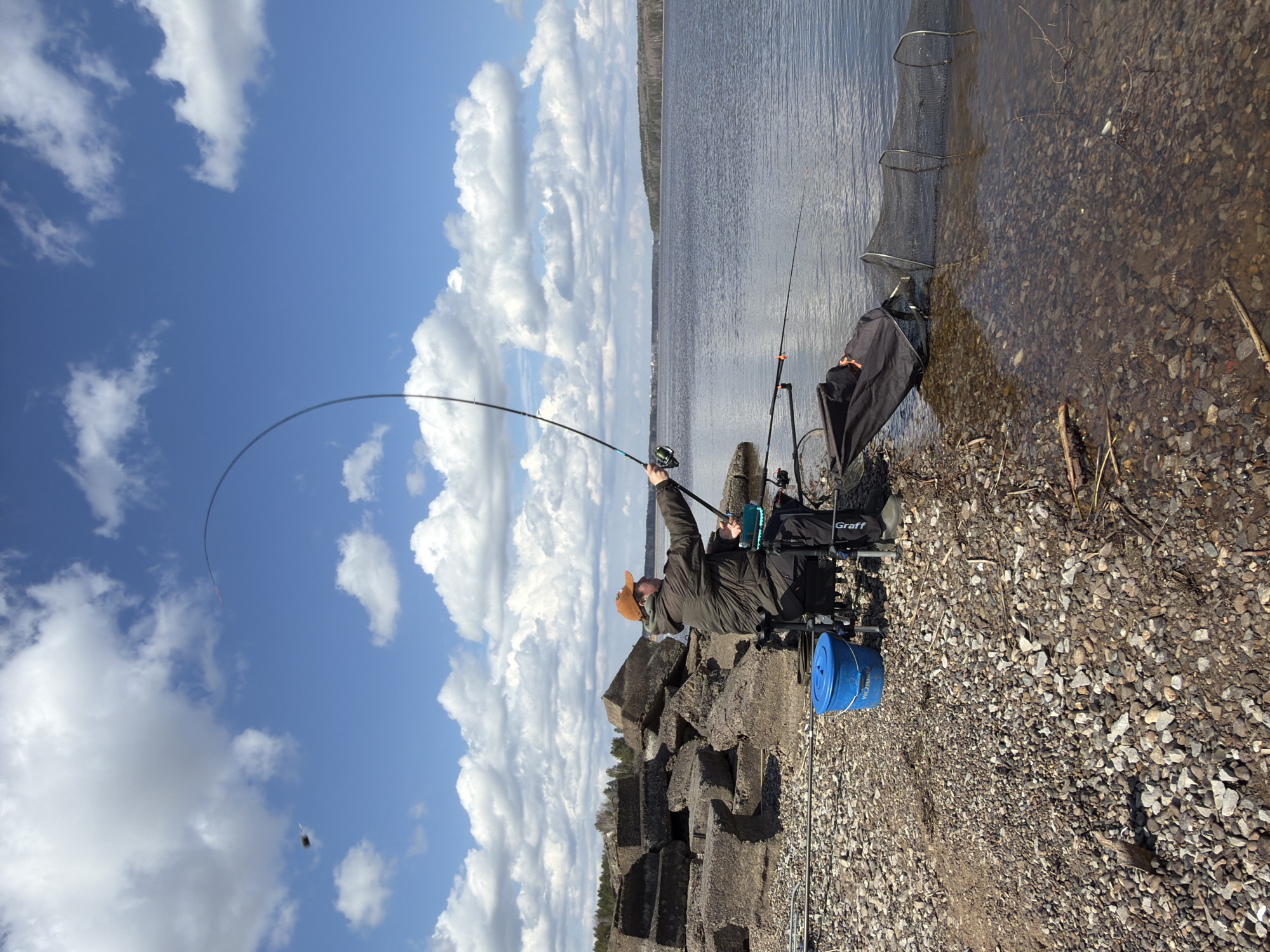 An outdoor, eye-level shot shows a man fishing on a sunny day on the rocky shore of a wide body of water, likely a river or lake. The sky is bright blue with numerous fluffy white cumulus clouds.
The man, wearing a dark jacket and a light brown hat, is seated on a fishing chair with a black fabric backrest. He is actively engaged in fishing, holding a long fishing rod with both hands. The rod is significantly bent, suggesting a fish might be on the line or he is in the process of casting or reeling in. To his right, another fishing rod is propped up vertically. A black keepnet, used for holding fish, is partially submerged in the clear, shallow water near the shore.
Behind the fisherman, several large, rough concrete blocks are visible, forming a low structure along the shore. A blue bucket, possibly a bait bucket, is placed on the gravel next to him. Text on the side of the bucket is partially visible, appearing to say "PRESTON INNOVATIONS." The brand "Graff" is visible in white lettering on the black bag or part of his fishing chair setup.
The shoreline is composed of small to medium-sized gravel and pebbles that transition into the water. Across the water, a distant treeline marks the horizon. The bright sunlight and scattered clouds suggest it is daytime, likely a pleasant day. The scene depicts a peaceful yet active fishing expedition.