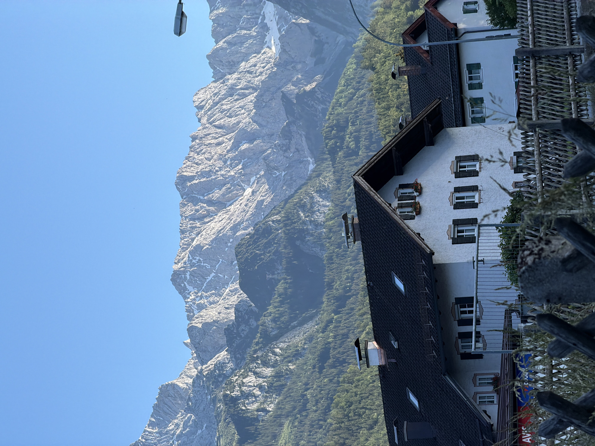 The image depicts a rural scene in Garmisch-Partenkirchen, Germany, with a focus on traditional Bavarian architecture and a prominent mountain range. In the foreground, two white houses with dark tiled roofs are visible. The house on the left has multiple windows with dark shutters and flower boxes, and several dormer windows on the roof. The house on the right also features windows with green shutters and a red-trimmed roofline. A wooden fence runs across the lower part of the image, with dry grass and foliage in front of it. Chimneys with protective caps are present on the roofs of both houses. In the background, a vast, jagged mountain range dominates the sky. The mountains are rocky with patches of snow, and their slopes are covered in dense green forest. The sky is a clear, bright blue. A lamppost is visible to the right of the mountains. The overall impression is one of a serene alpine village setting.