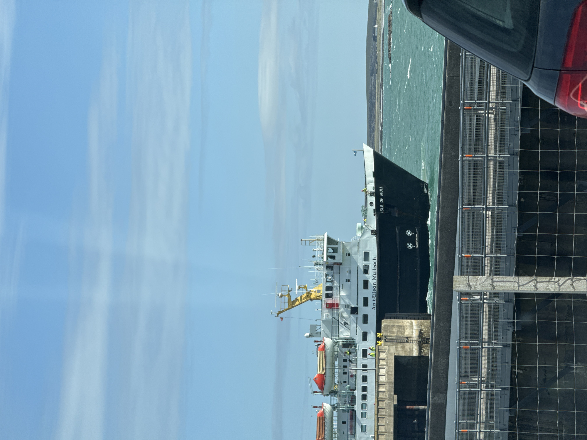 The image shows a ferry, "Isle of Mull", docked next to a concrete pier. The sky is partly cloudy. A section of the stern of a gray car is visible on the right side of the image. A wire fence is in the foreground. The location is unknown.