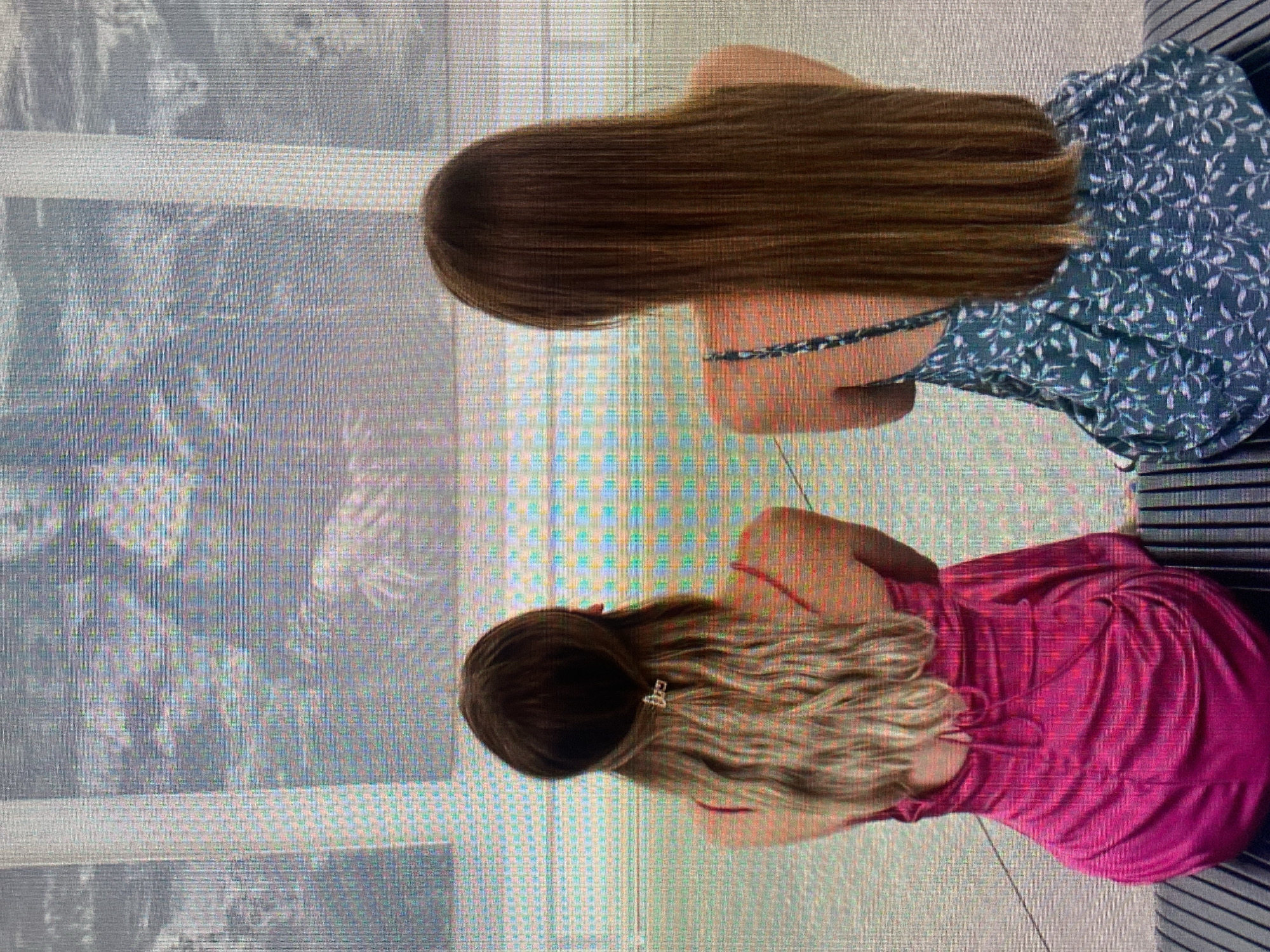 Two women are seated side-by-side. One has long brown hair and wears a blue floral print dress. The other has long blonde hair with a hair clip and wears a pink dress. They are seated in front of a window or display case which reflects a muted image. The location is likely in Łódź, Poland, but there is no visible visual verification of this information.