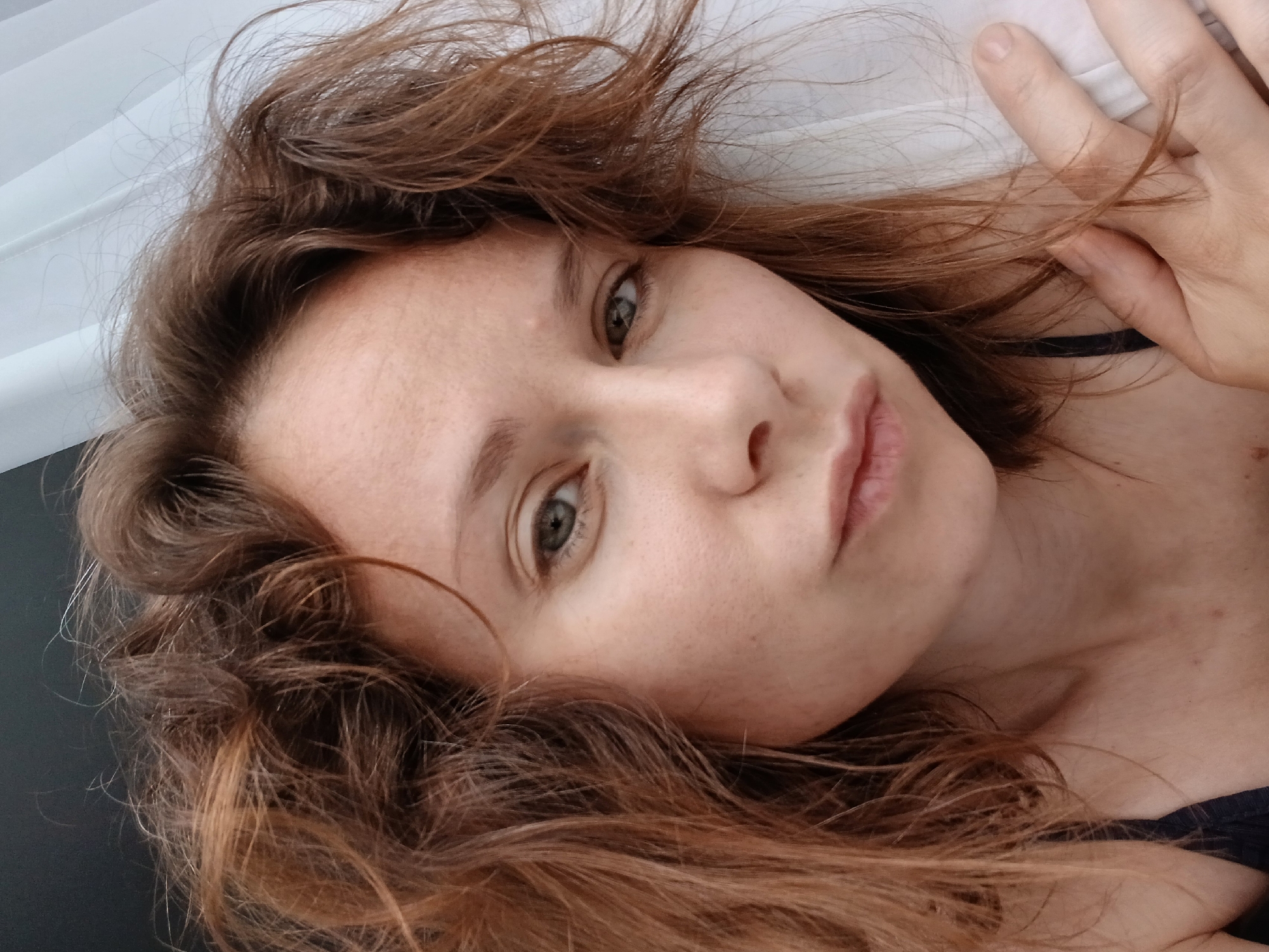 A woman with reddish-brown, messy hair, fair skin with freckles, and light green eyes looks directly at the camera. Her lips are slightly pursed. She is wearing a dark blue strap top. Her right hand is resting on her chest, near her shoulder, partially obscuring white fabric behind her. The background is dark on the left and white and textured on the right, suggesting indoor lighting with a window or sheer curtain. The setting does not provide specific Yekaterinburg landmarks.