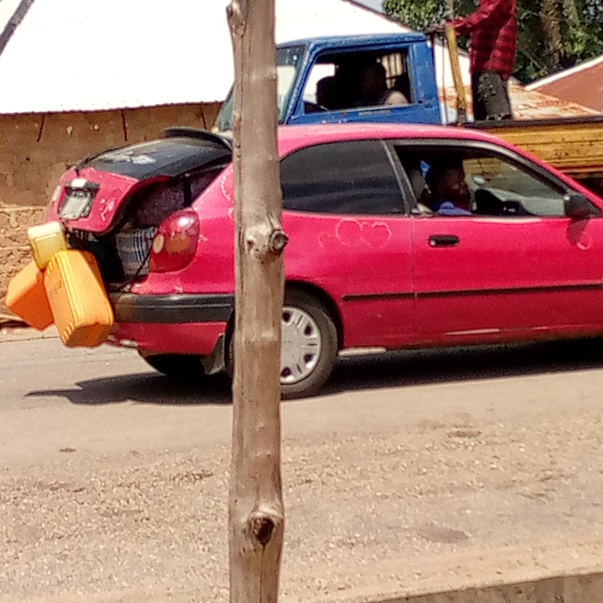 The image shows a red Toyota Corolla hatchback parked on a road in Mararaba, Nigeria. The car's trunk is open, and several yellow jerrycans are sticking out. A man is driving the car. In the background is a blue pickup truck with two people in the cab. A third person is standing in the truck bed. The setting appears to be an urban or semi-urban environment. The road surface is damaged. A tree trunk is partially obscuring the view.