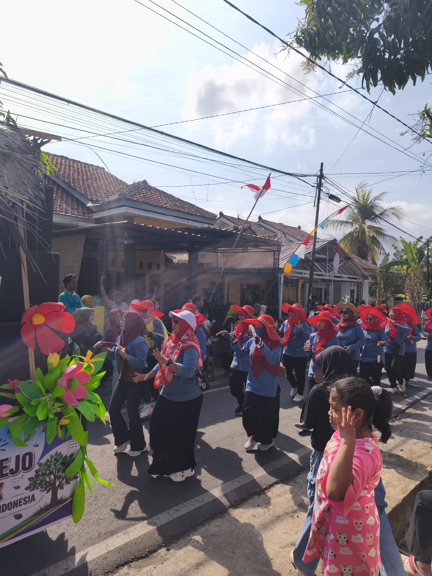 In the city of Kutoarjo, Indonesia, a parade is in progress on a sunlit street. A group of women, dressed in blue shirts and long black skirts, march together, many wearing bright red wide-brimmed hats and red headscarves. They appear to be in motion, possibly dancing or performing. To the left of the frame, a large, colorful float is visible, adorned with oversized red and pink flowers and green leaves. The float has a banner with text, including the word "KUTOARJO". Behind the participants, houses with tiled roofs line the street, and utility poles with numerous wires crisscross the sky. The sky is partly cloudy with bright sunlight casting shadows on the asphalt road. On the right side of the image, a young girl in a pink dress with an owl print watches the parade.