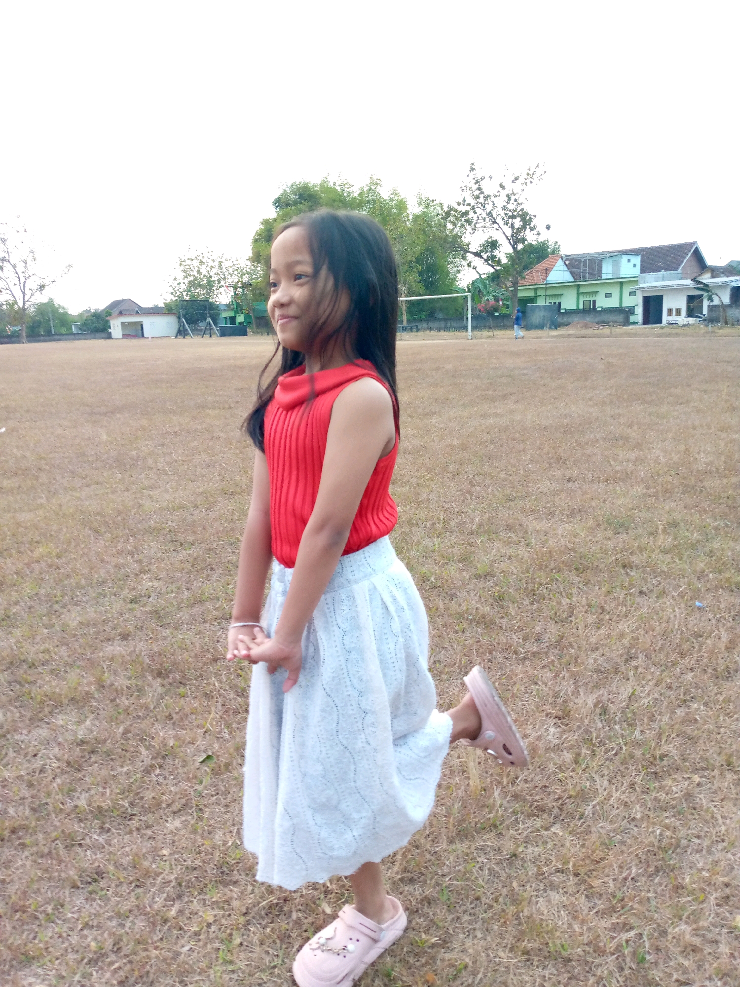 A young girl with long, dark hair stands in a dry, grassy field. She is wearing a red, sleeveless top with a high neckline and a light-colored skirt. Her left foot is raised and bent at the knee, and she is looking to her left. Her hands are clasped together in front of her.
In the background, there is a soccer goal, trees, and buildings. The sky is overcast, suggesting it could be morning or late afternoon. The overall scene appears to be in a rural or semi-urban area in Indonesia. The girl's posture and gaze suggest she might be posing for a picture or about to move. There is no visible text in the image.