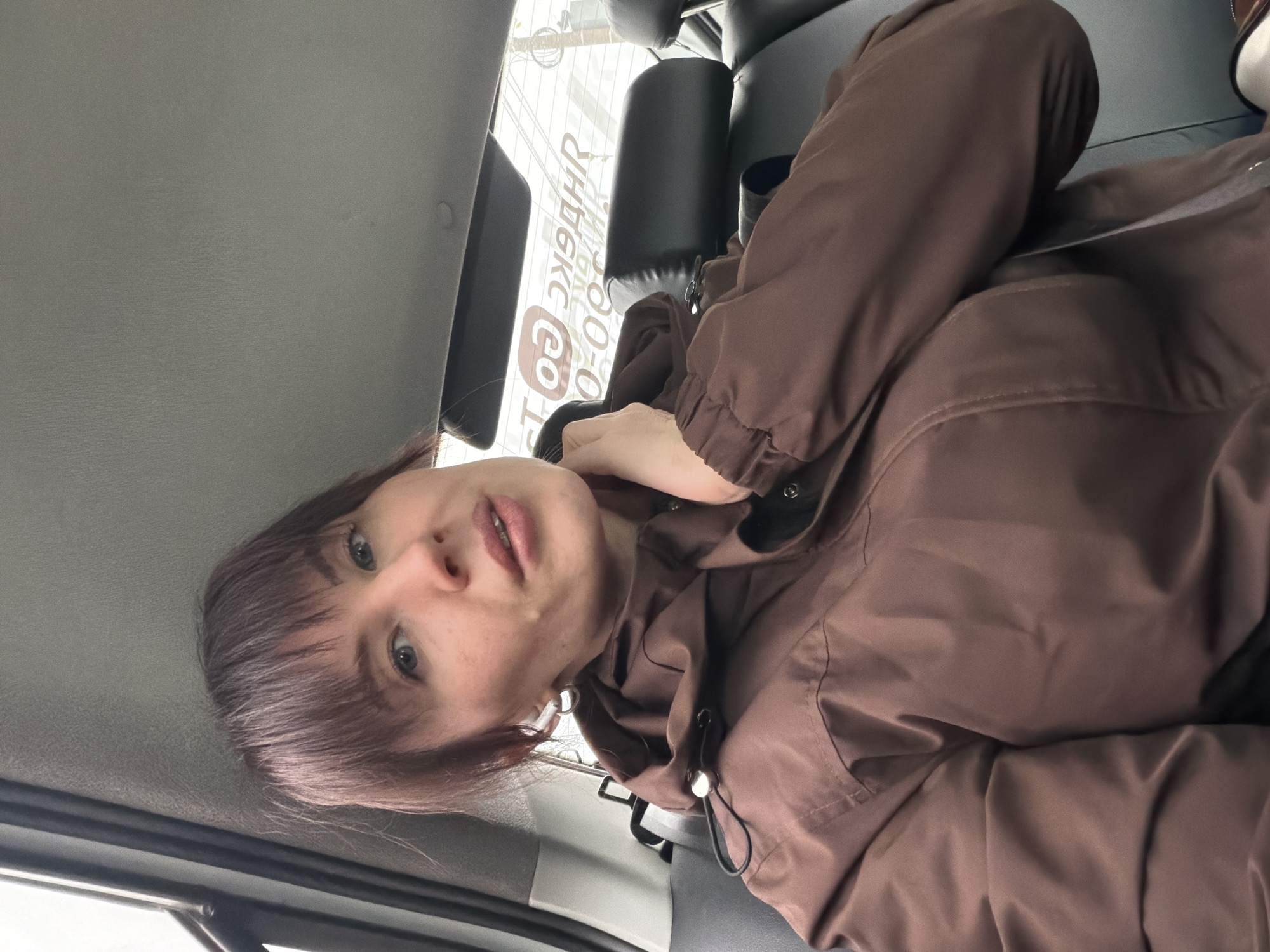 A woman with shoulder-length brown hair and bangs, wearing a brown hooded jacket with a seatbelt across her chest, sits in a vehicle. The interior of the vehicle is gray. Outside the vehicle window, a sign appears partially visible and readable in reverse as "Perm" (Пермь) in Russian. This indicates the location of the image is the city of Perm, Russia.