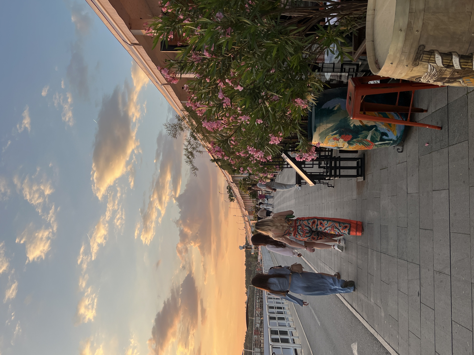 The image depicts a sunset scene in Belgrade, Serbia, on a waterfront promenade. In the foreground, three young women are walking away from the camera on a paved walkway. The woman on the left wears a light blue denim set, the middle woman wears a white top and skirt, and the woman on the right is in a vibrant, patterned jumpsuit. They are walking past a row of buildings with awnings, adorned with flowering oleander bushes. To the right of the walkway, decorative barrels and a red stool are visible. In the background, a river or waterway is present, with a white railing separating it from the promenade. The sky is illuminated by the warm hues of the setting sun, with scattered clouds. Other people are visible further down the promenade, suggesting a public gathering place.