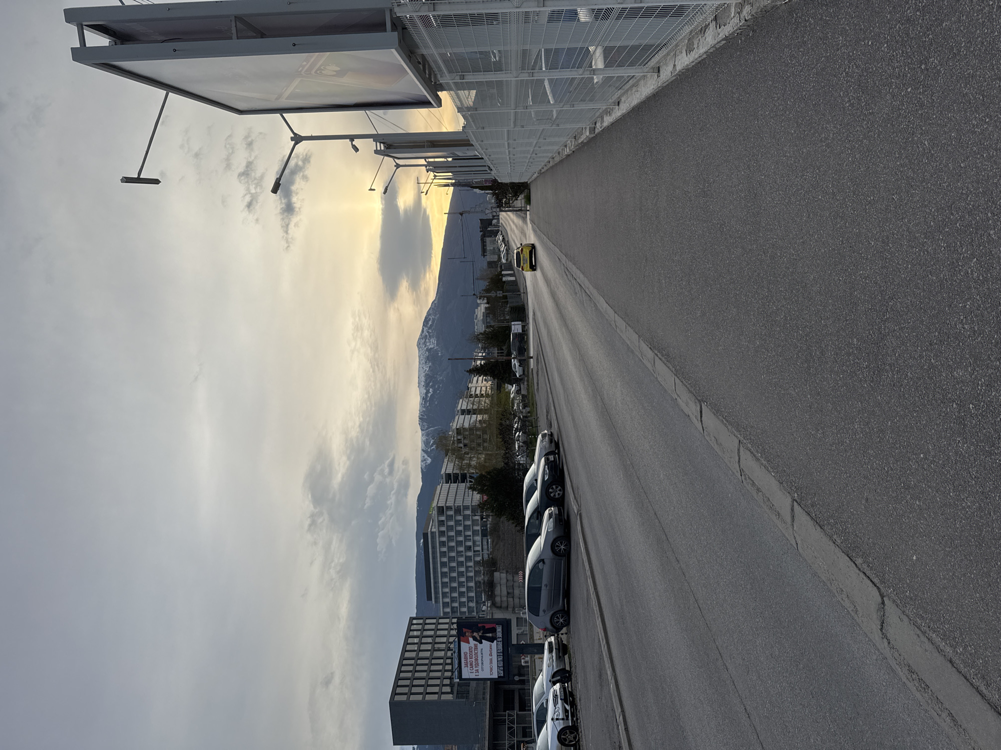 This image shows a street in Sofia, Bulgaria, lined with parked cars on the right and buildings and mountains in the background. The perspective is angled, with the road and sidewalk stretching into the distance. The sky is cloudy with a hint of sunlight breaking through. A large billboard with text in Bulgarian is visible on the side of a building, and a mountain range is seen in the far background. A yellow car is driving on the road in the direction opposite to the camera position.