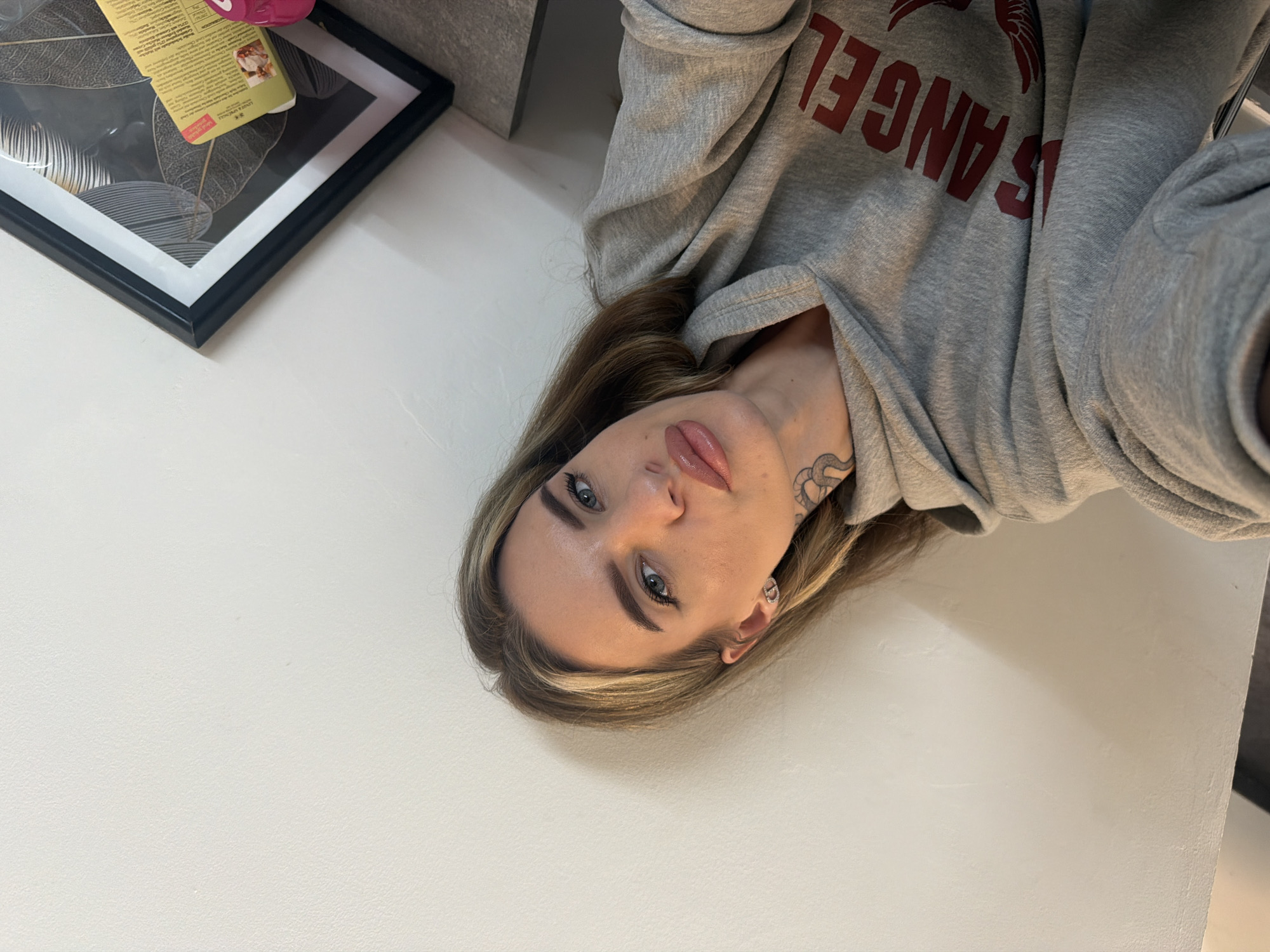 A young woman is depicted in a casual, direct portrait, likely a selfie, lying on a light-colored surface in an indoor setting. She has long, light brown hair with blonde highlights, blue-grey eyes, and pink, glossy lips. Her eyebrows are well-defined, and she wears mascara. A coiled, black and white tattoo, possibly a snake, is visible on her neck/upper shoulder, and she has a small, intricate silver-colored earring in her visible ear.
She is dressed in a grey hooded sweatshirt (hoodie) featuring large red text on the front. Part of the text is clearly visible as "LOS ANGEL," strongly suggesting "LOS ANGELES."
In the upper left background, behind her head, a black-framed picture with abstract black and white botanical art, depicting large leaves, is visible. Resting on top of this frame is a small, rectangular box or container with some text on its label, and a small pink object is placed nearby. The overall lighting is soft and even, indicative of indoor ambient light. The scene, set in Berlin, Germany, appears to be a personal and relaxed moment.
