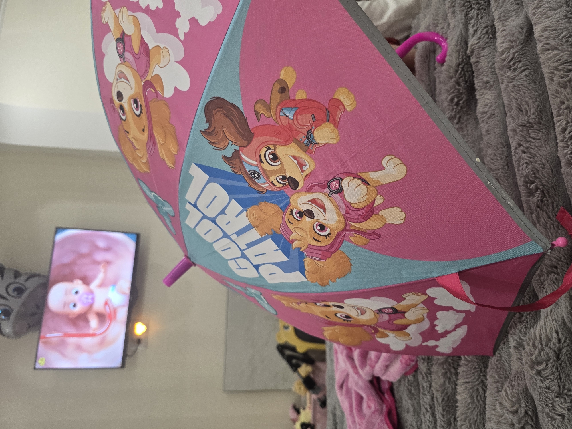 In an indoor setting, a pink and teal umbrella with cartoon dogs and the words "COOL PATROL" is prominently displayed on a gray, furry surface, likely a bed or blanket. The umbrella features animated characters from Paw Patrol. To the left and slightly behind the umbrella, a television screen displays a cartoon image of a baby in a womb with a pacifier. Above the television, a gray, cat-shaped decoration is attached to the wall. The environment appears to be a child's room or a recreational space.