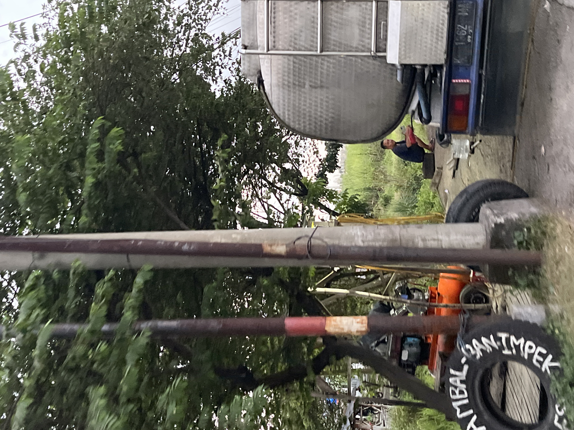 The image captures a roadside scene in Pamotan, Indonesia. In the foreground, a large tire with white lettering that appears to read "AMBAL SANTI MPEK" is visible. Behind it, an orange industrial machine, possibly an air compressor, is situated. A person wearing a dark blue shirt and red shorts is squatting near the rear of a large silver tank truck. The truck has a blue cab and a license plate displaying "K 162 WH" and a date stamp of "09.23". The environment is overgrown with green grass and trees, suggesting a rural or semi-rural setting. Two utility poles are prominent in the mid-ground, with one exhibiting red and white markings. The lighting indicates it is daytime, possibly late afternoon due to the soft light.