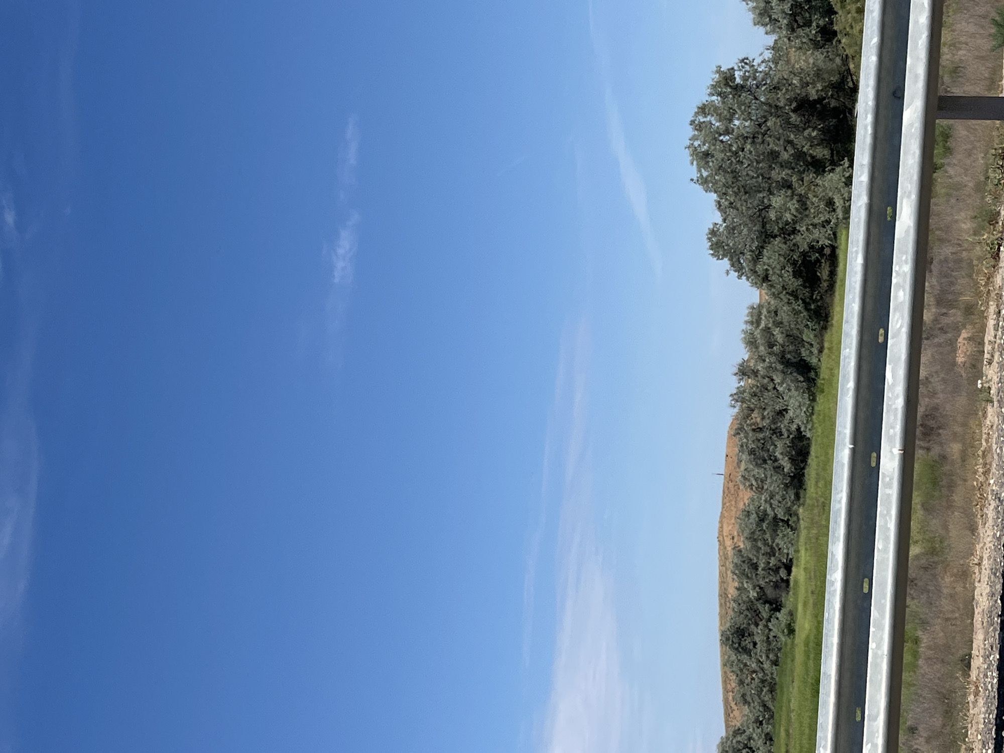 The image shows a clear blue sky with a few wispy clouds. In the middle ground, there is a rolling hill covered in dry, brownish vegetation, with a line of green grass and trees in front of it. A metal guardrail runs across the foreground of the image, obscuring some of the lower landscape. The scene appears to be a rural or roadside landscape in the vicinity of Bakhtemir, Russia. There are no people or discernible actions taking place.