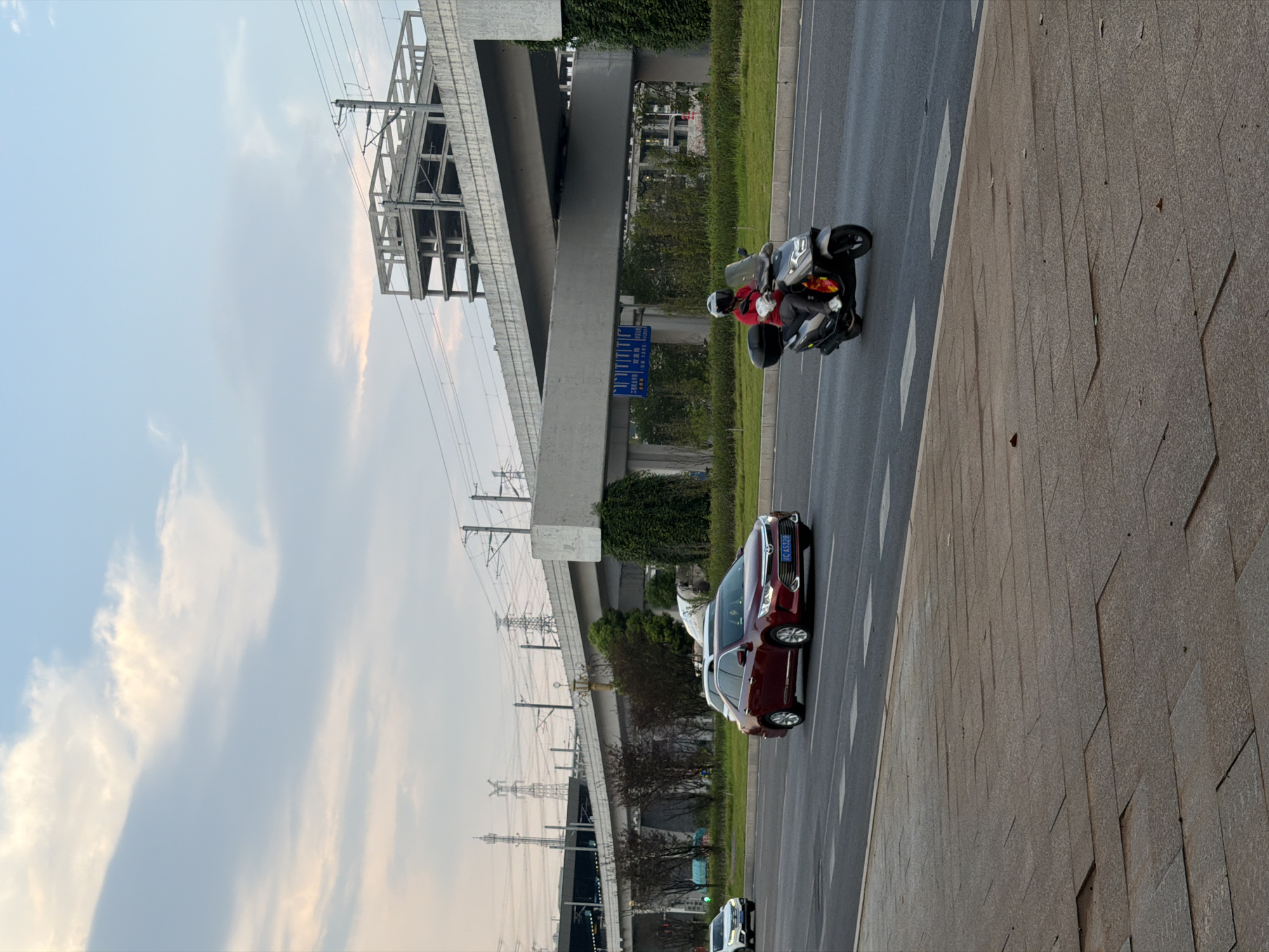 In Jiangjin, China, a motorcyclist wearing a red shirt and white helmet rides a dark gray scooter on an asphalt road. The scooter has a black storage box on the rear. To the left of the scooter, a red sedan with a license plate reading "C AS328" is also traveling on the road. In the background, a multi-level elevated structure, likely for transportation, is under construction, with concrete beams and supports visible. Power lines and transmission towers are also present in the distance. The sky is cloudy with patches of light blue and a hint of orange at the horizon, suggesting it might be late afternoon or early evening. A sidewalk made of rectangular stone tiles is in the foreground.