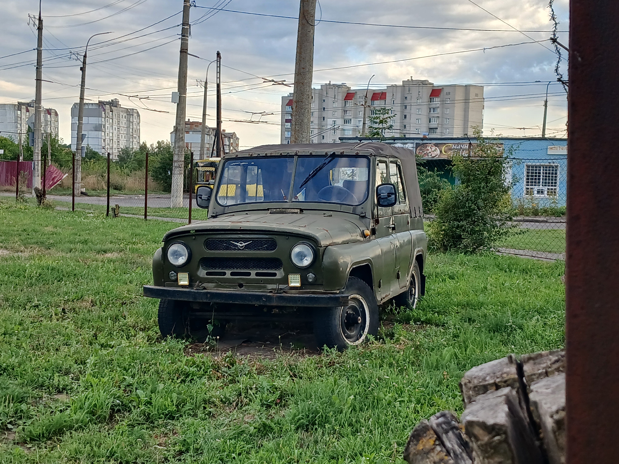 A vintage, olive-drab UAZ-469 off-road vehicle is parked in a grassy, overgrown lot. The vehicle is positioned front-center in the frame, facing slightly to the right. The UAZ has a soft top, a metal bumper, round headlights, and a grille with the UAZ logo. There are power lines and poles in the background, along with several multi-story apartment buildings. A blue building with a white-gridded window is visible to the right, behind a chain-link fence. The foreground shows a blurry section of rough-hewn wood. The scene is under a cloudy sky.