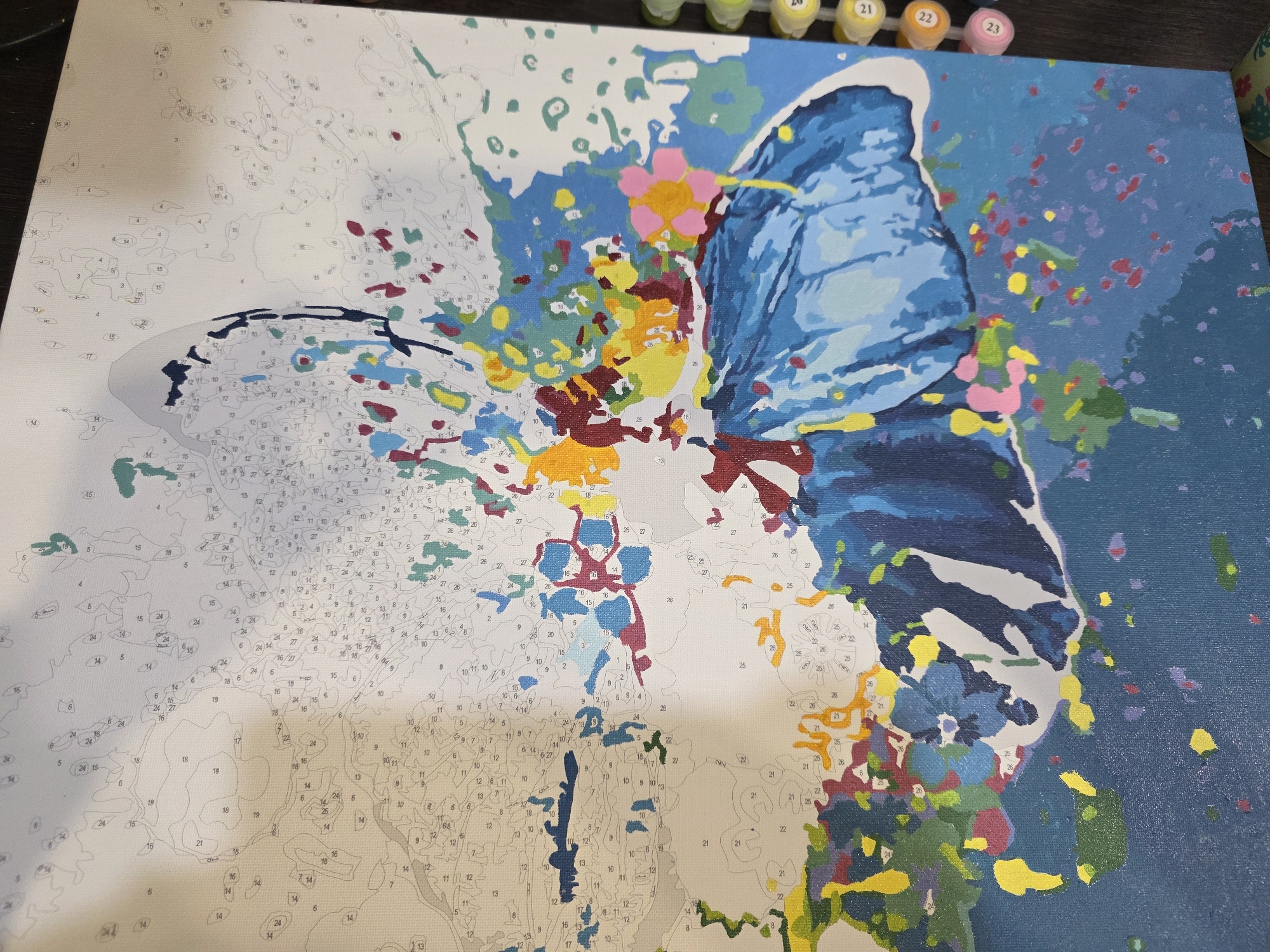 This image captures a close-up, high-angle view of a partially completed paint-by-numbers canvas, set in Chaikovsky, Russia. The canvas features a vibrant, abstract design centered around a butterfly. The right side of the canvas shows a large blue butterfly wing, almost fully painted with various shades of blue and white highlights. Surrounding the butterfly are abstract splatters and shapes in a medley of colors including bright yellow, pink, red, light blue, green, and dark blue, indicating a dynamic, colorful background.
The left side and bottom sections of the canvas remain unpainted, revealing the intricate outlines of numerous small areas, each marked with a specific number. These numbers, such as 3, 4, 5, 6, 7, 8, 9, 10, 11, 12, 13, 14, 15, 16, 17, 18, 19, 20, 21, 22, 23, 24, 25, 26, and 27, guide the painter on which color to use for each segment. In the upper right corner, several small pots of paint are visible, corresponding to the numbers on the canvas. Labels on the paint pots show numbers 20, 21, 22, 23, and 24, indicating the specific colors ready for application.
The activity depicted is the ongoing process of painting, showcasing the contrast between the unpainted, numbered canvas and the brightly colored, completed sections. The canvas rests on a dark surface, possibly a table or desk. The image highlights the detailed work involved in paint-by-numbers, transforming a numbered outline into a colorful piece of art.