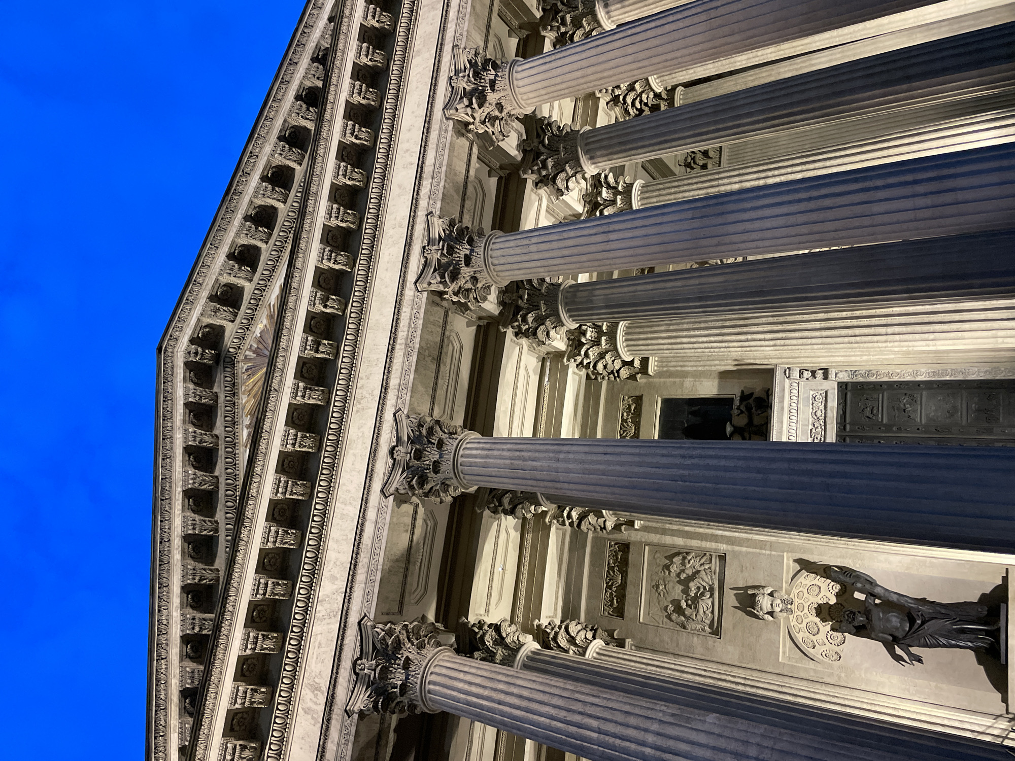This is a low-angle, vertically oriented shot of a large, ornate building with classical architecture. The building features massive fluted columns with detailed Corinthian capitals. Above the columns, a section of the building's facade is visible, adorned with intricate carvings and decorative elements. A bright blue sky fills the upper left portion of the frame, indicating it's likely dusk or dawn, or possibly a very clear day. The architectural details suggest a grand public building, possibly a museum, courthouse, or government institution. The lighting suggests artificial illumination on the building, highlighting the sculptures and architectural features. No people are visible in the frame. The scene evokes a sense of grandeur and historical significance.