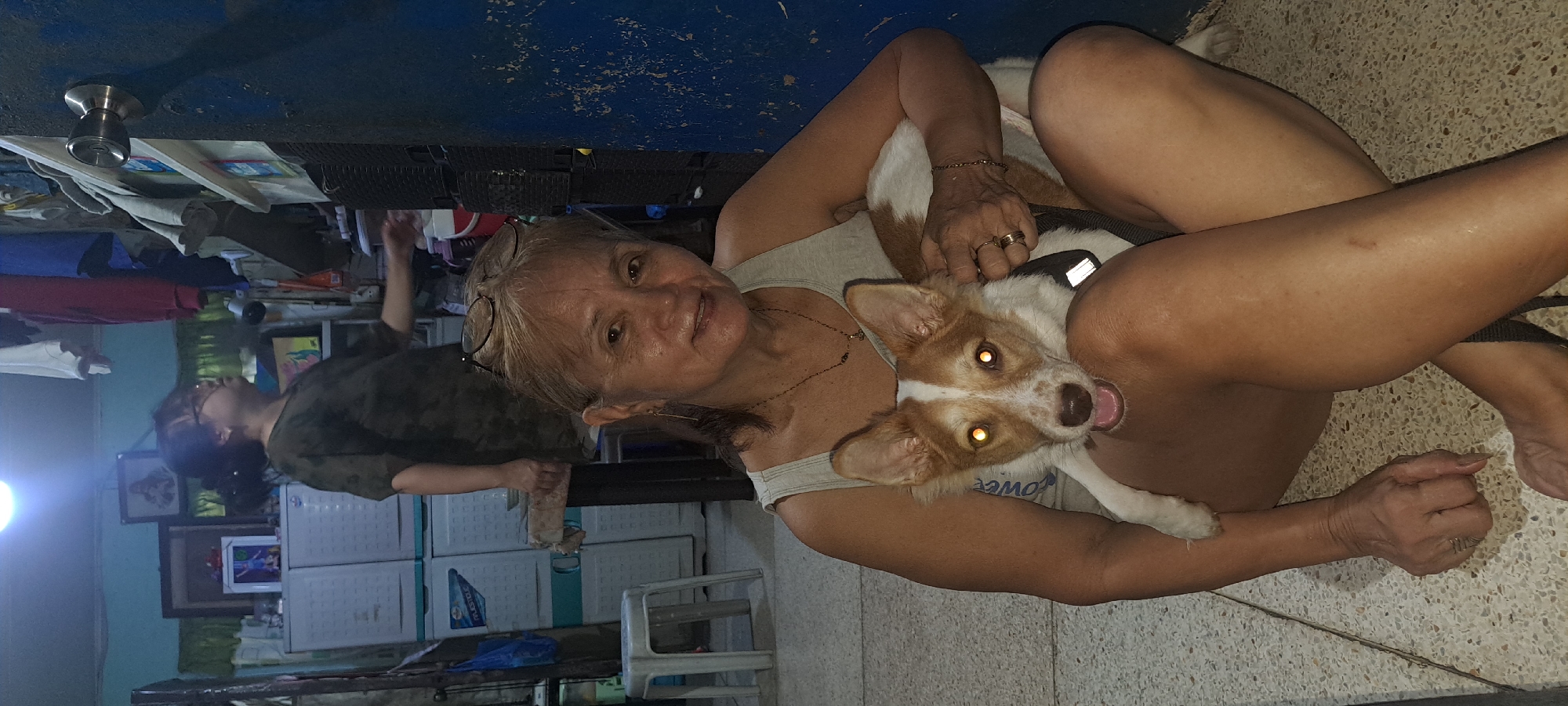 In a dimly lit room in Mandaluyong, Philippines, a woman with blonde hair and glasses perched on her head sits on a terrazzo floor, cradling a medium-sized dog with white and brown fur. The dog has bright, glowing eyes and its mouth is slightly open. The woman is wearing a sleeveless top and a necklace. In the background, another person is visible, standing and looking away from the camera. The room appears cluttered with various objects, including white storage cabinets, clothing hanging from a rack, and a doorway on the right side of the frame. The overall impression is of a casual, indoor setting.