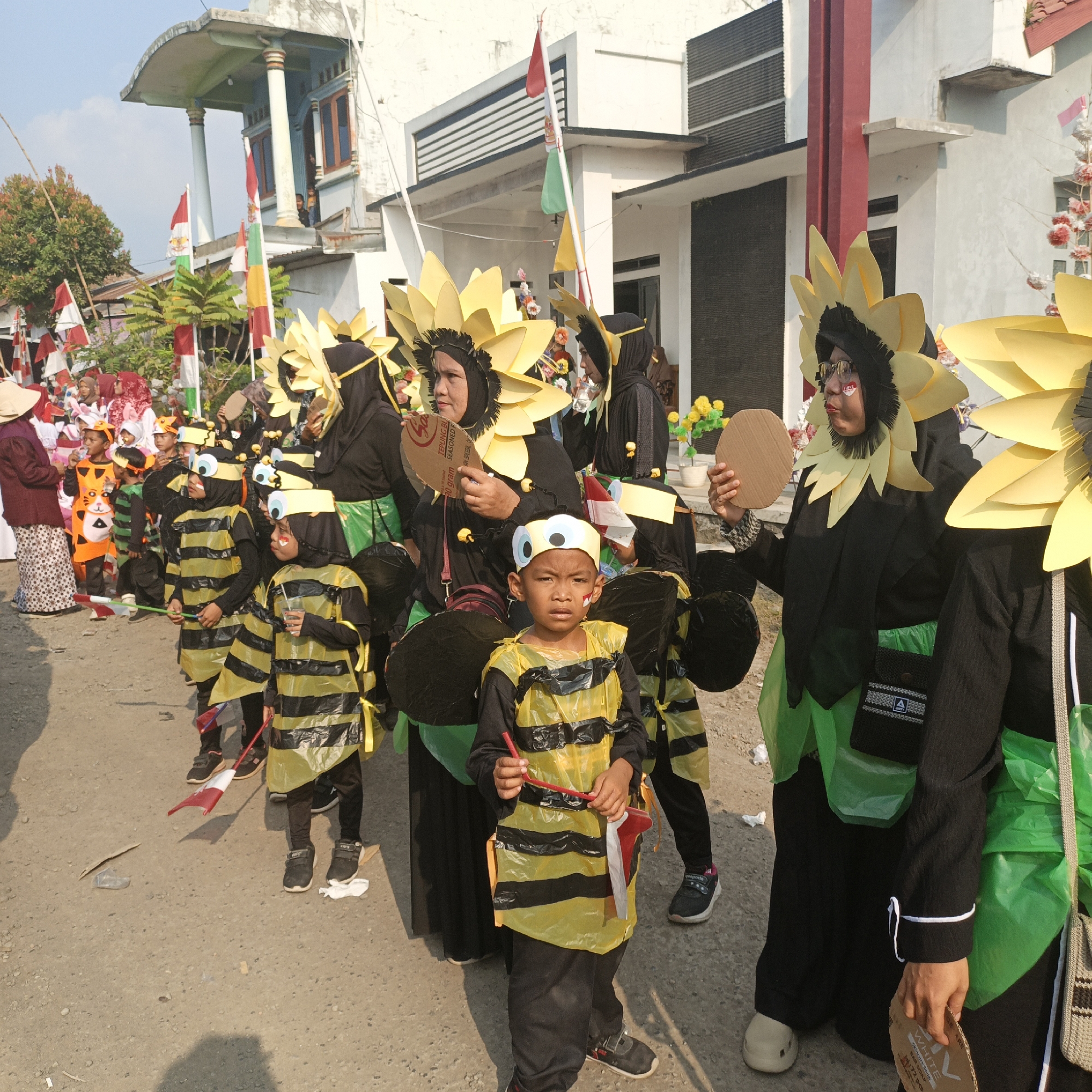 In the city of Tanahbaya, Indonesia, a group of children and adults participate in a parade, likely celebrating a national holiday, given the presence of Indonesian flags and the festive attire. The individuals are dressed in elaborate costumes. Several children are adorned in black and yellow striped bee costumes, complete with antennae headbands and wings. Other participants, possibly adults, wear large, yellow sunflower headdresses and black robes, holding circular cardboard props. The procession walks along a dusty, unpaved road, with a building featuring columns and multiple flags in the background. The scene is illuminated by bright daylight, suggesting it is outdoors during the day.