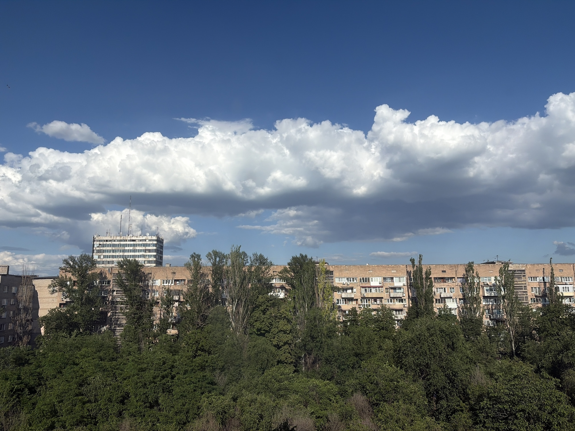 This media file shows a panoramic view of a residential area on a sunny day. The scene is dominated by a clear blue sky with large, fluffy white clouds. In the foreground, a dense canopy of green trees stretches across the frame, obscuring the lower parts of the buildings.
Behind the trees, several multi-story apartment buildings are visible. The buildings are constructed with brick and concrete, featuring numerous windows and balconies. One taller building, possibly an office or administrative building, stands out in the background, topped with communication equipment. The architecture suggests a Soviet-era urban landscape.
The time of day appears to be daytime, likely mid-morning or early afternoon, given the bright sunlight casting shadows and illuminating the clouds. The weather is pleasant, characterized by a blue sky and scattered clouds. There are no discernible people or specific activities taking place, creating a static and serene urban scene. No text is visible in the image.