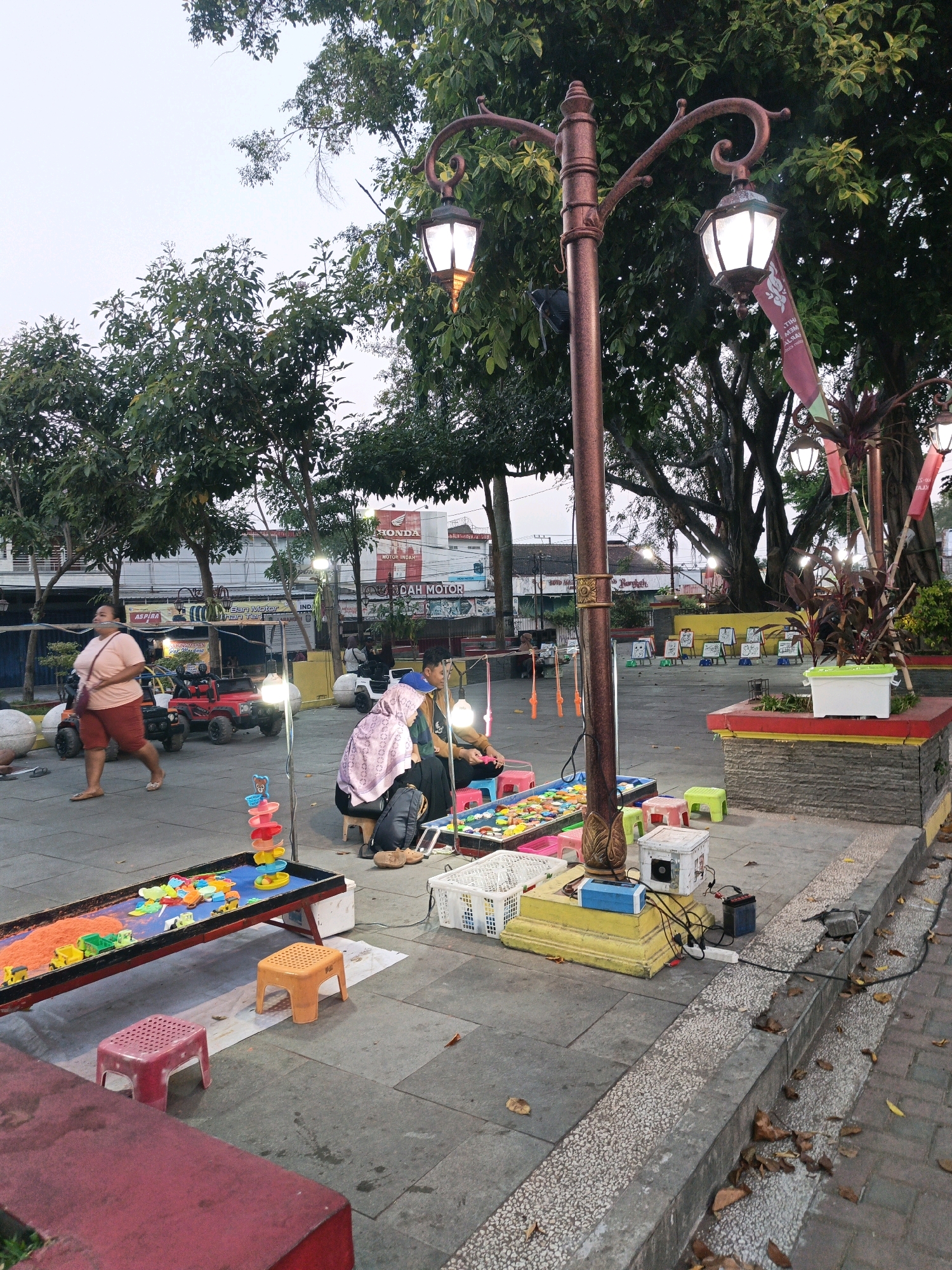 This image depicts a street scene in Klaten, Indonesia, likely in a public square or park area. In the foreground, a sandpit with toy vehicles is set up, surrounded by small plastic stools of various colors. A lamppost with decorative elements stands prominently. In the mid-ground, two individuals are seated near the sandpit, possibly attending to it or interacting with a customer. A woman in a light-colored shirt and orange shorts is walking away from the camera. Several small, toy electric cars are parked nearby, suggesting a children's play area. Further in the background, buildings with signage, including "HONDA," are visible, indicating commercial activity. Trees line the area, providing shade. The ground is paved with stone tiles. The overall atmosphere suggests a relaxed, outdoor public space with recreational activities available.