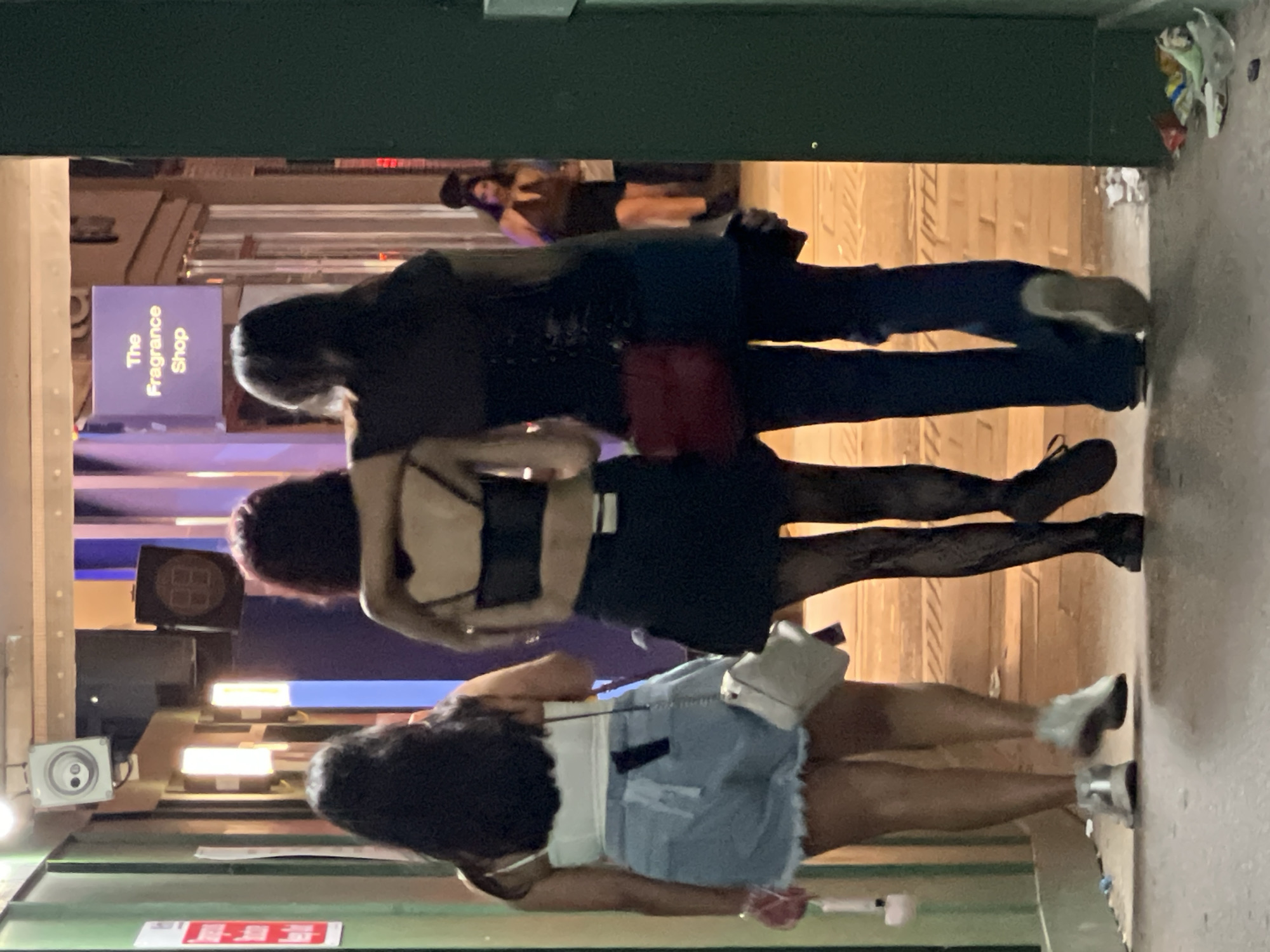Three women are standing together in front of a shop in London, United Kingdom. The shop sign to the left reads "The Fragrance Shop." The first woman is wearing a black top and blue jeans. The second woman is wearing a black top and black shorts with tights. The third woman is wearing a white top and denim shorts. The background shows buildings and lights typical of an urban street scene. The ground is concrete, and there is debris and trash near the wall in the upper right corner.