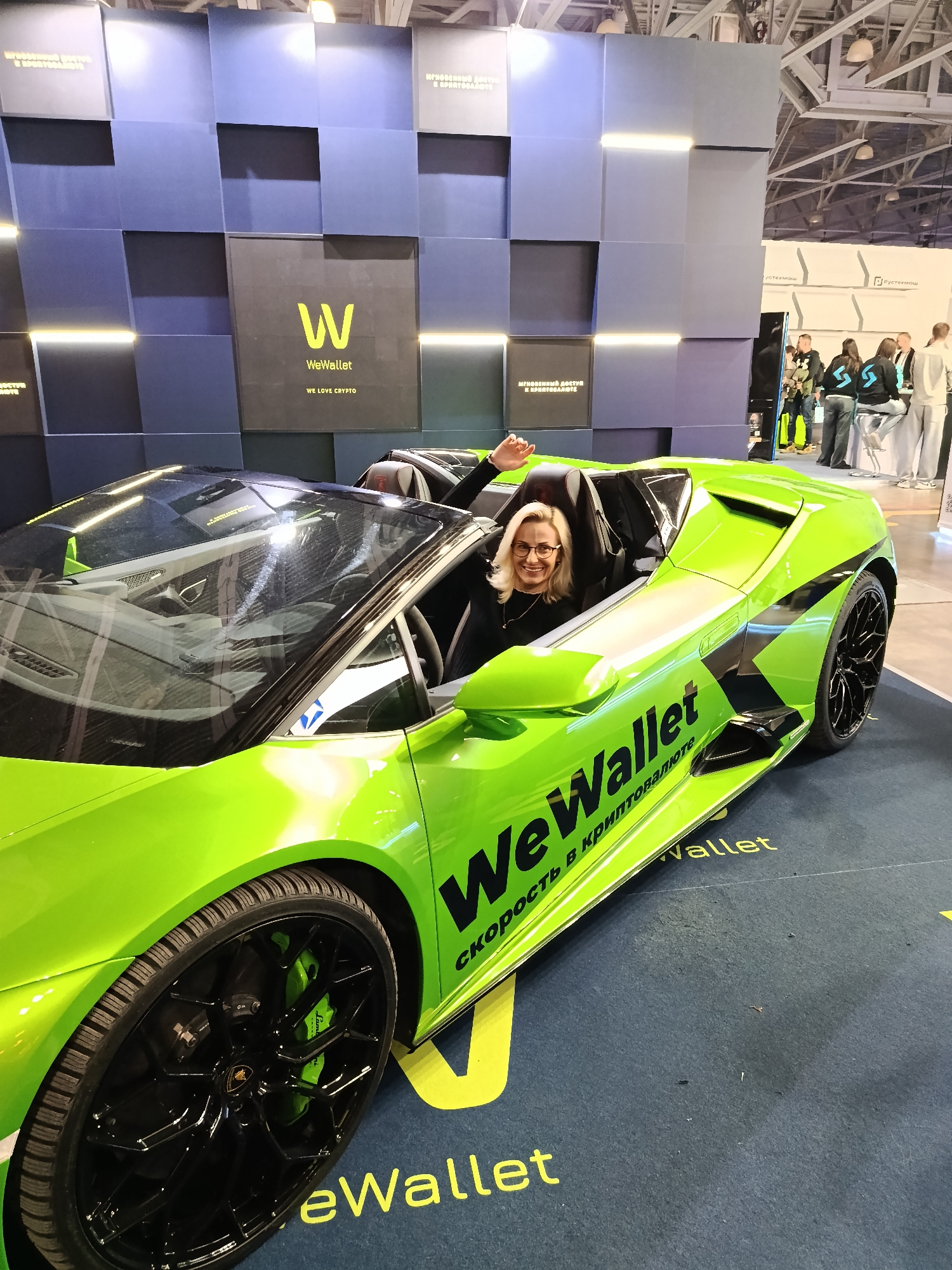 The image shows a lime green Lamborghini sports car with the words "WeWallet" printed on its side. The car is parked indoors, possibly at an exhibition or convention, with a blue and gray textured wall in the background featuring the WeWallet logo and branding. A woman with blonde hair and glasses is sitting inside the car, waving. Other people are seen in the background, some standing and some seated. There is text in Russian on the wall behind the car that translates to "Instant access to crypto" and on the car itself, "Speed in cryptocurrency". Given the use of Russian text and brands like "Рустехмаш", the location is Moscow, Russia.