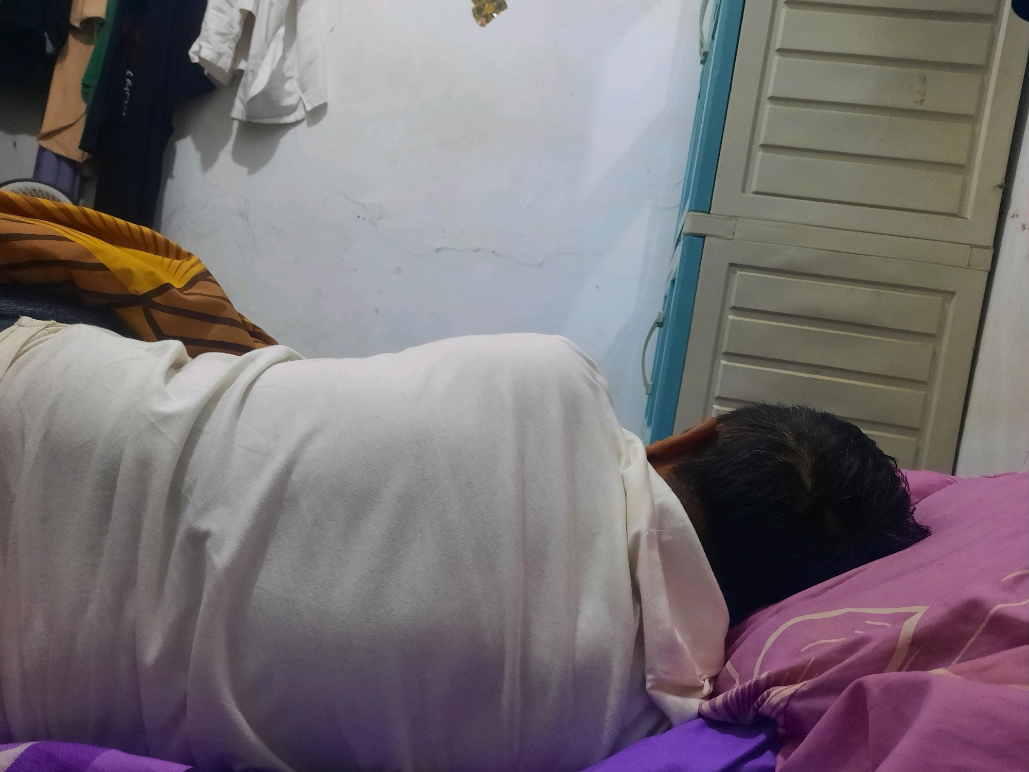 An adult male is lying on a bed in a room, sleeping. He is wearing a white t-shirt and is covered by a blanket with a yellow and brown striped pattern. The bed has a pink and purple patterned sheet. In the background, there is a white wall with some clothing hanging on it, and a light-colored wardrobe with blue trim. The scene appears to be in a residential setting, likely a bedroom. The location is Nagrak, Indonesia.
