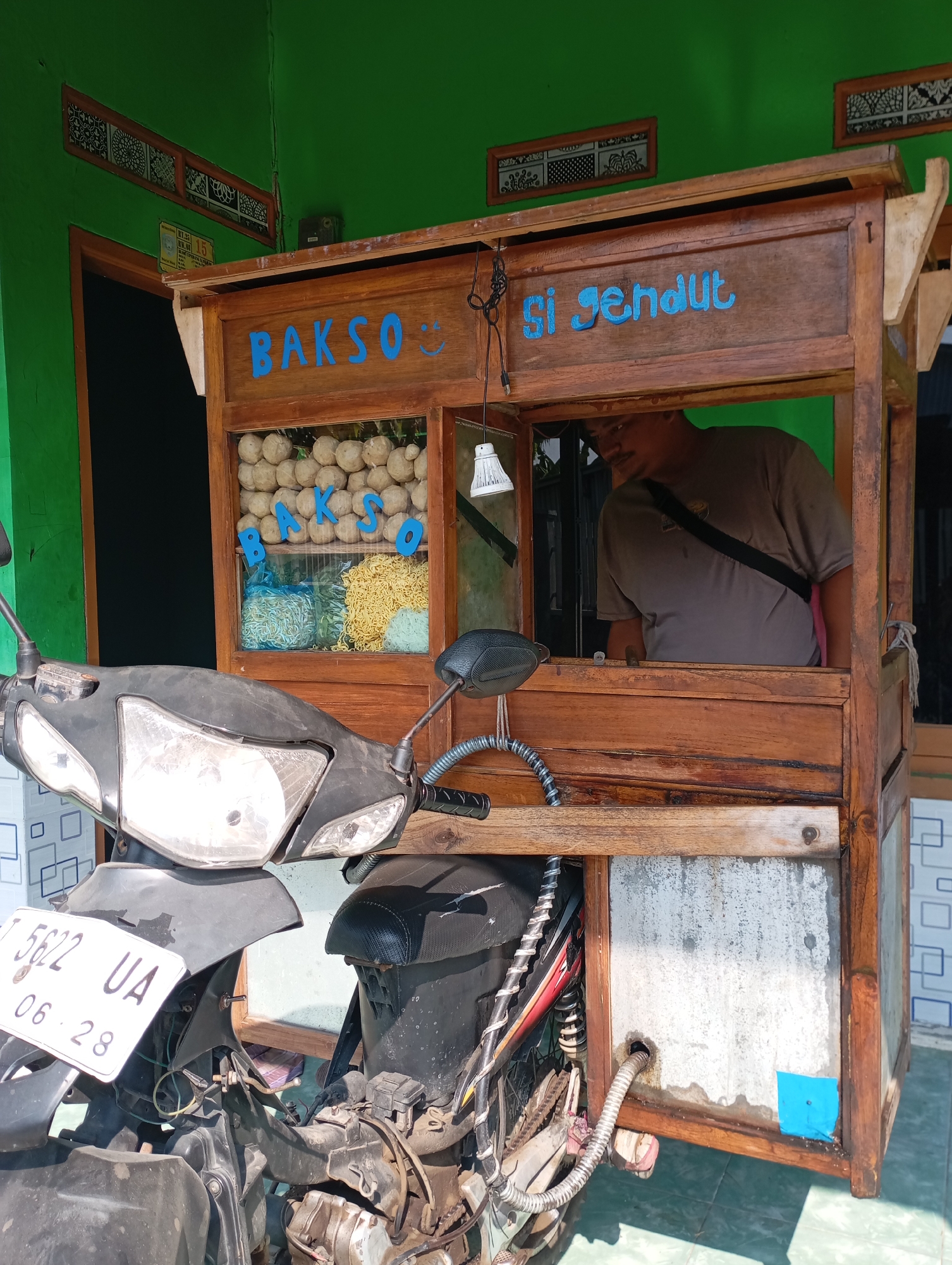 In the city of Kecamatan Haurgeulis, Indonesia, a wooden food cart is parked next to a motorcycle. The food cart, which appears to be a mobile bakso (meatball) stand, has a sign in blue letters that reads "Bakso Si Gendut". The cart displays a variety of ingredients, including meatballs, noodles, and what appears to be green vegetables. A man, likely the vendor, is visible inside the cart. The motorcycle is parked in front of the cart, its license plate reading "B 5622 UA" with a date of "06.28". The overall scene suggests a typical street food vendor setup in a residential or semi-commercial area.
