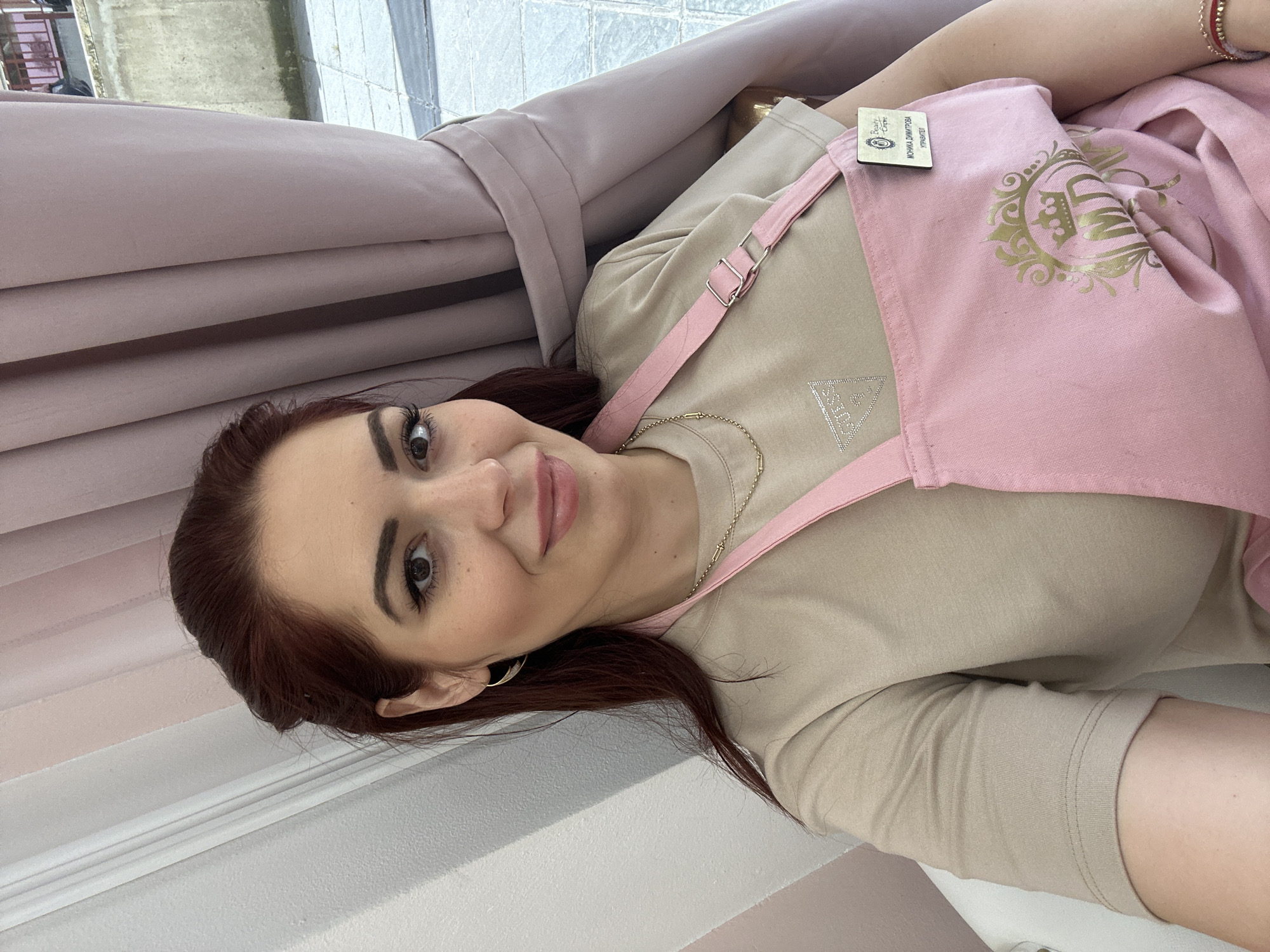 The image depicts a woman with auburn hair, wearing a beige t-shirt and a pink apron with a gold crown emblem, possibly in a commercial setting. The backdrop includes pink curtains and a window showing an exterior view, likely in a building. Sofia, Bulgaria is the assumed location.