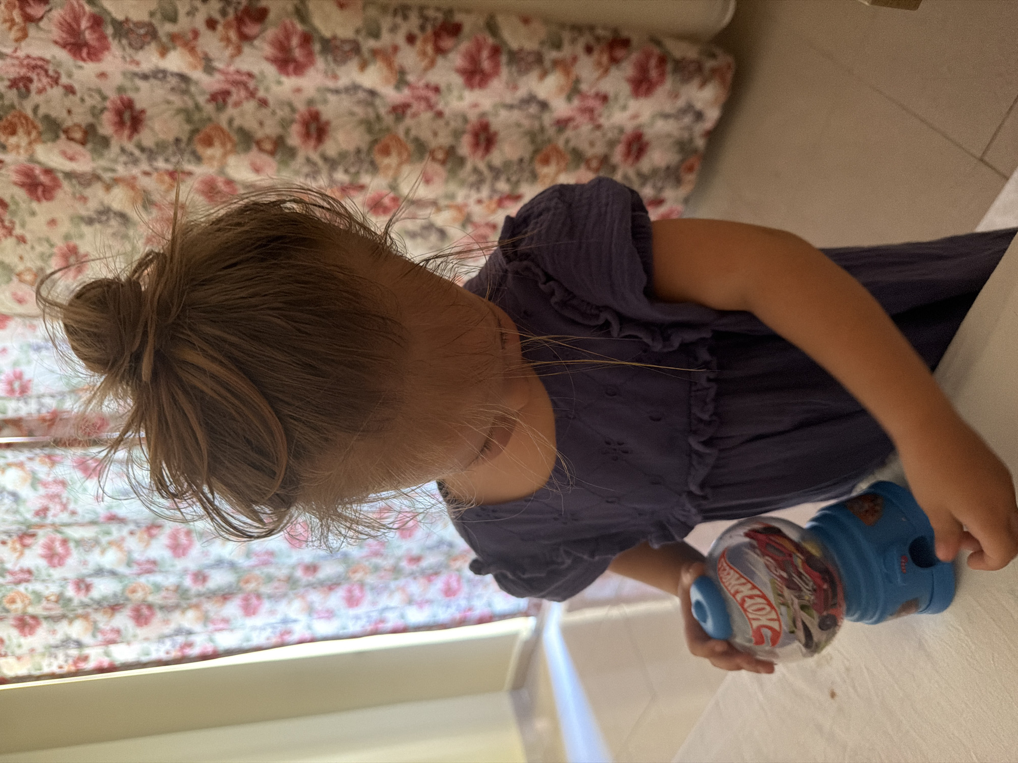 A young girl with her hair in a bun is standing at a table, holding a blue gumball machine. The gumball machine is clear and contains Hot Wheels cars. She is wearing a dark blue dress. In the background, there is a floral curtain. The scene appears to be indoors, possibly in a home in Fethiye, Turkey.