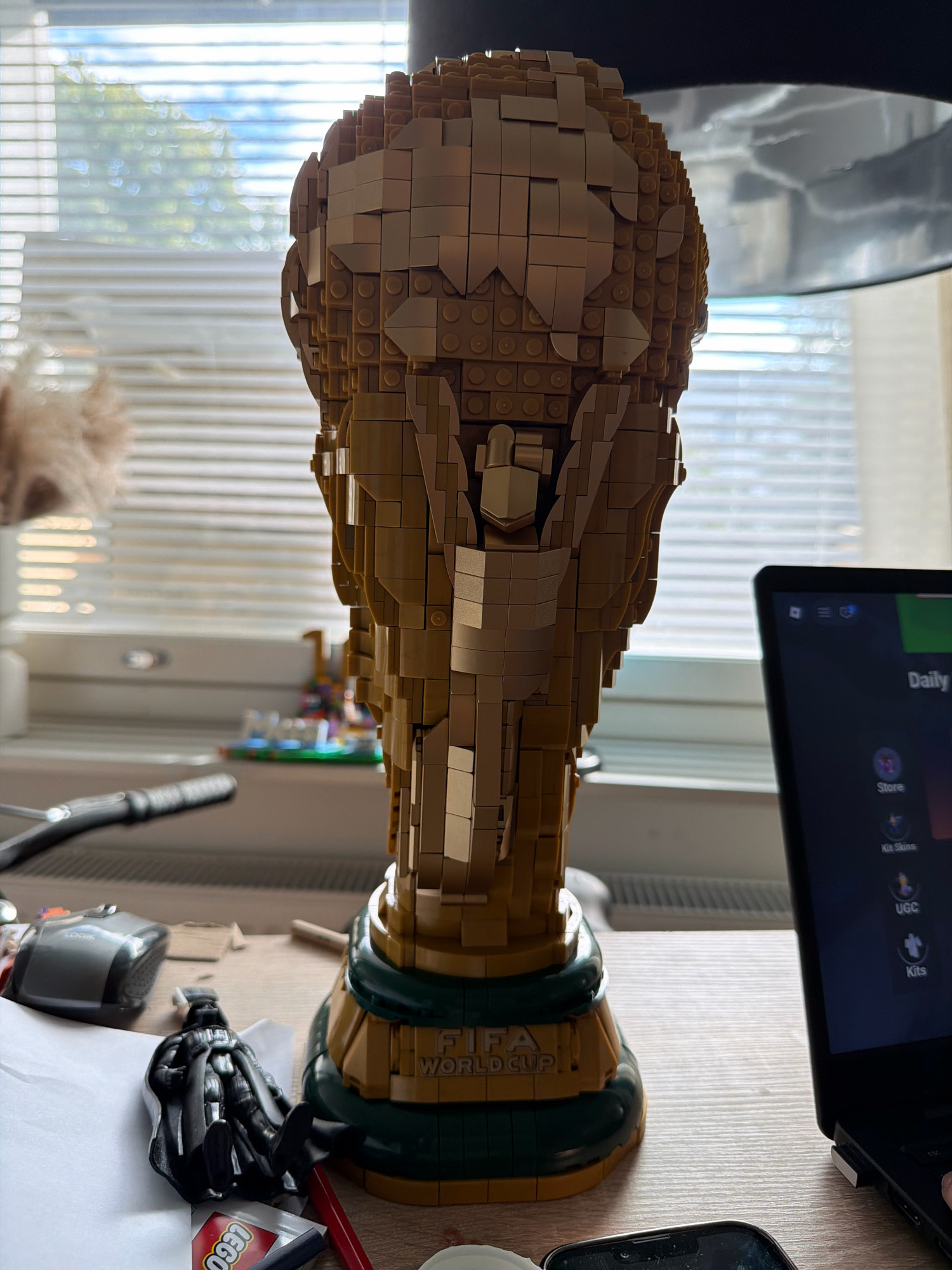 The image depicts a desk scene with a large LEGO model of the FIFA World Cup trophy as the central subject. The trophy is made of tan-colored bricks and has a green base with "FIFA WORLD CUP" printed on it. To the left of the trophy, there is a computer mouse and a black action figure, possibly Darth Vader. A portion of a laptop screen is visible on the right, displaying what appears to be a gaming or app interface with text like "Daily," "Store," "Kit Skins," and "UGC." The background shows blinds on a window, with trees and sky visible through them. The desk surface is made of wood-textured material. The scene is indoors, likely a room or office, with no specific identifying landmarks for Helsinki, Finland, or any people present.
