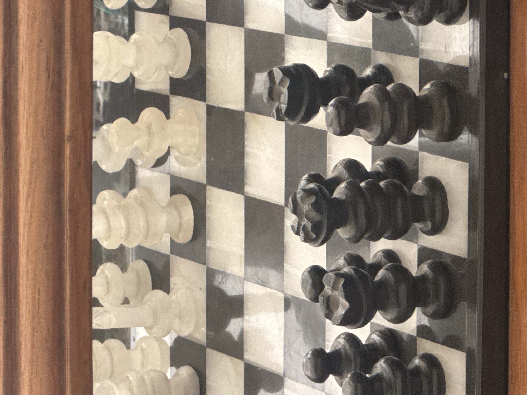 The image shows a vertically oriented, close-up view of a chessboard with chess pieces. The board has a black and white checkered pattern, and the pieces are arranged in the initial setup position. The white pieces, located on the left side of the image, are translucent. The black pieces, on the right, are solid black. The chessboard appears to be part of a decorative or display piece, framed by wood on the left and right edges of the image. The exact location in Puebla, Mexico, cannot be determined from the image alone.